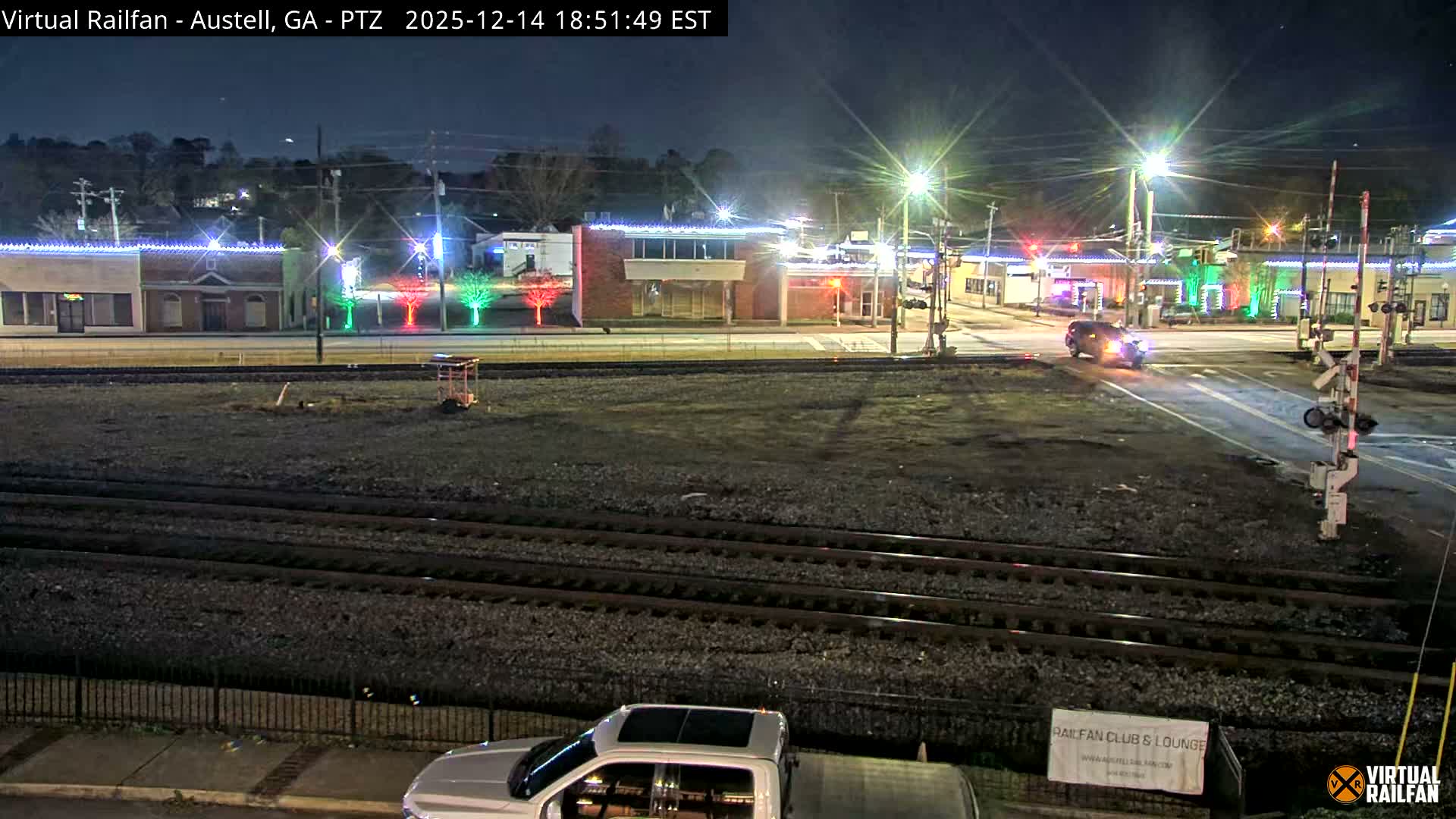 A rainy night scene overlooks multiple railroad tracks and a glistening town street intersection, where buildings are adorned with white string lights and colorful red and green tree lights, illuminated by streetlights and a car's headlights.