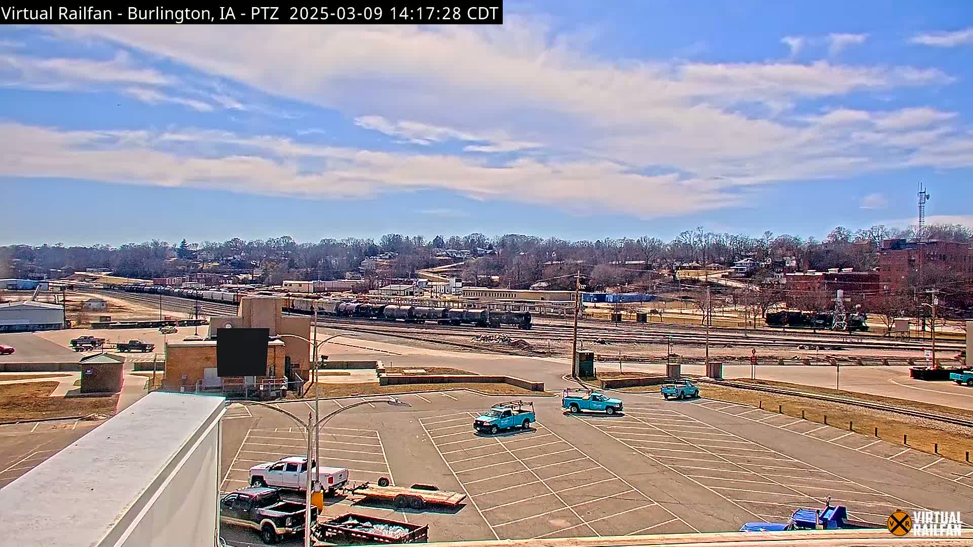 A mostly sunny day reveals a train yard with several freight cars, a parking lot with work trucks, and a treeline in the background.