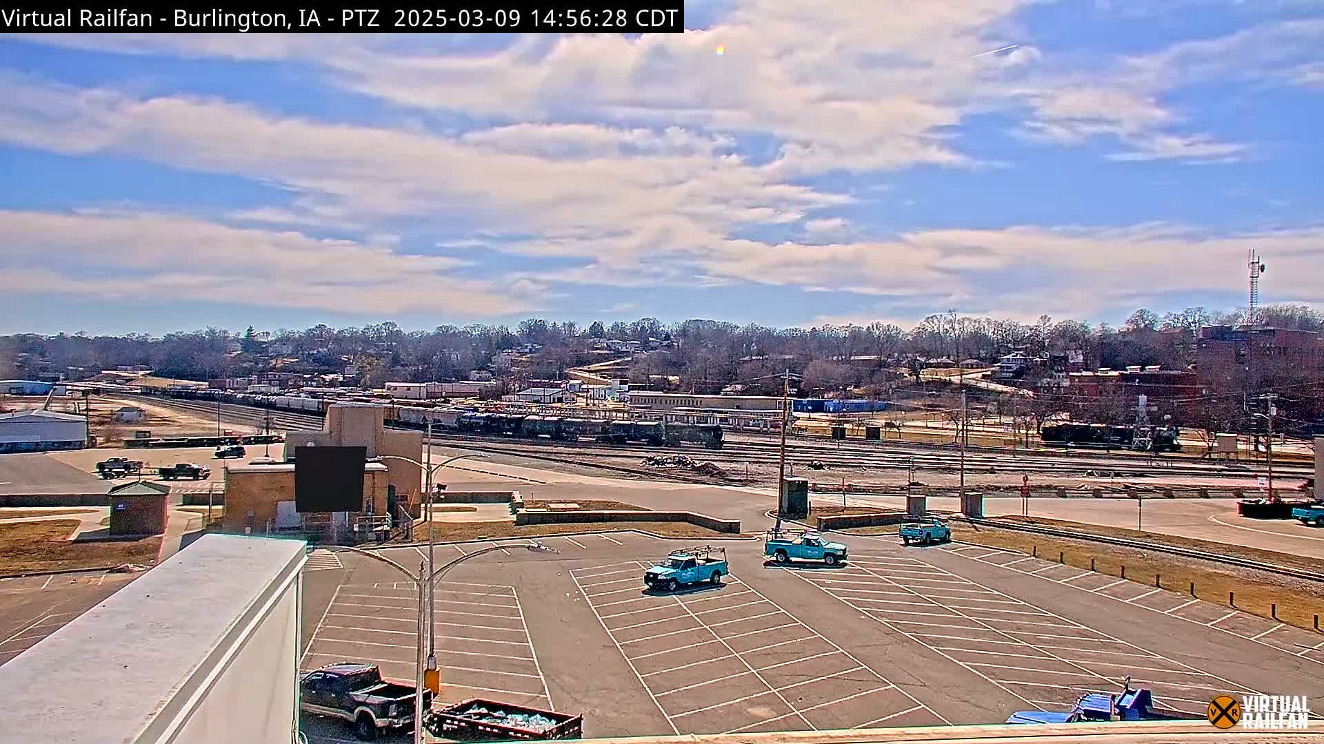 A mostly sunny day reveals a train yard with several railcars, a parking lot with several trucks, and a treeline in the background.