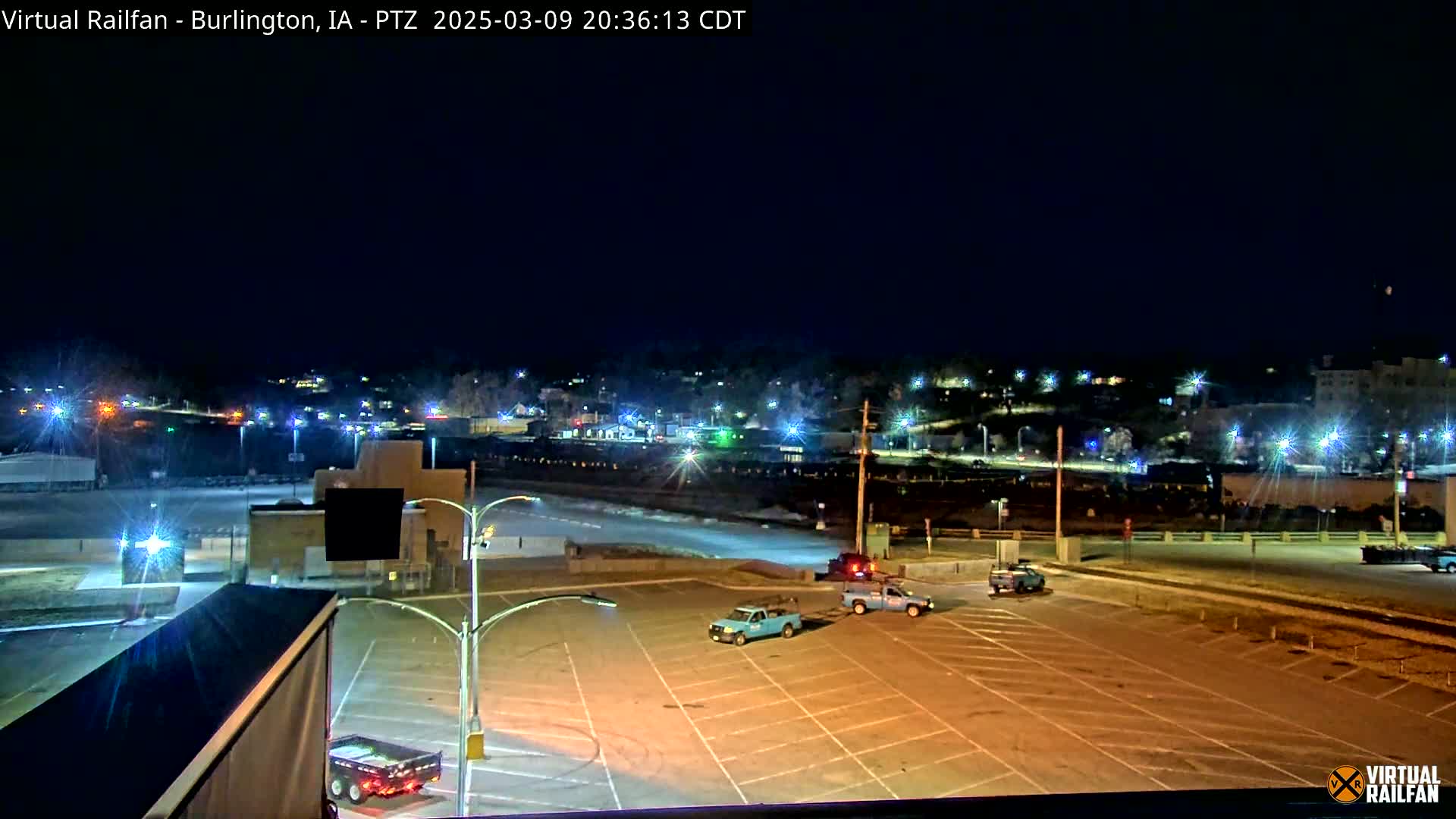 A nighttime, brightly lit view of an empty parking lot with a few vehicles and a town in the background under a clear sky.