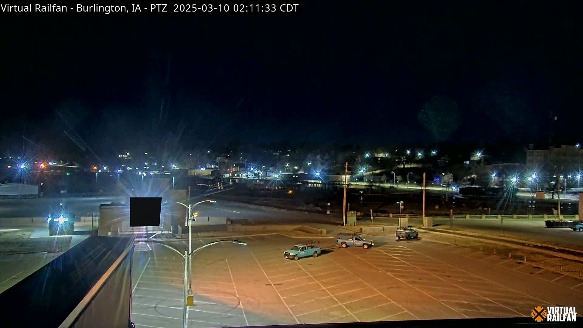 A mostly empty parking lot at night is illuminated by streetlights, with a few vehicles present and a distant cityscape visible under a clear, dark sky.
