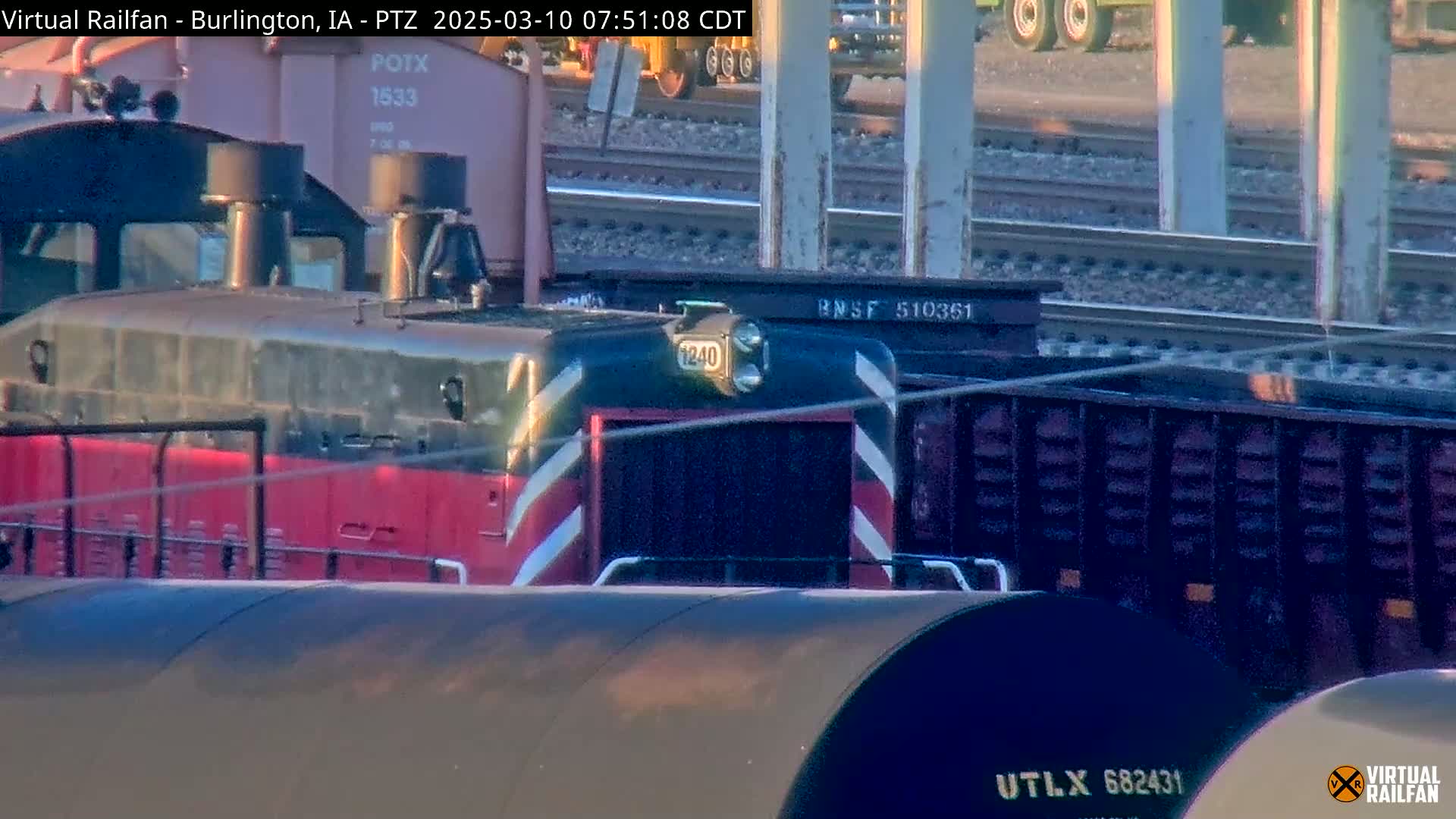 A red and black locomotive is partially visible behind several freight cars under a partly sunny sky.