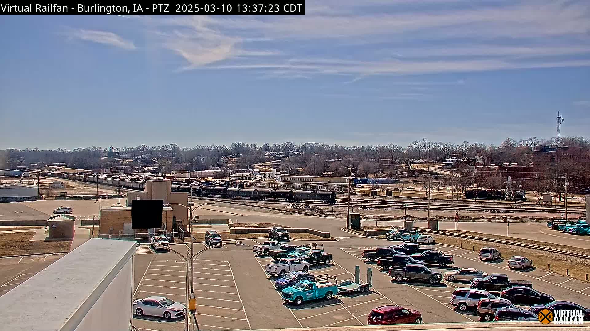 A mostly sunny day reveals a train yard with a long freight train and a nearby parking lot full of various vehicles; in the distance are buildings and bare trees.