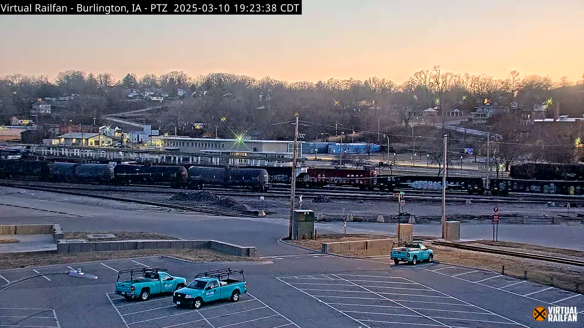 Two teal pickup trucks are parked in an empty lot overlooking a train yard with numerous freight cars under a twilight sky.