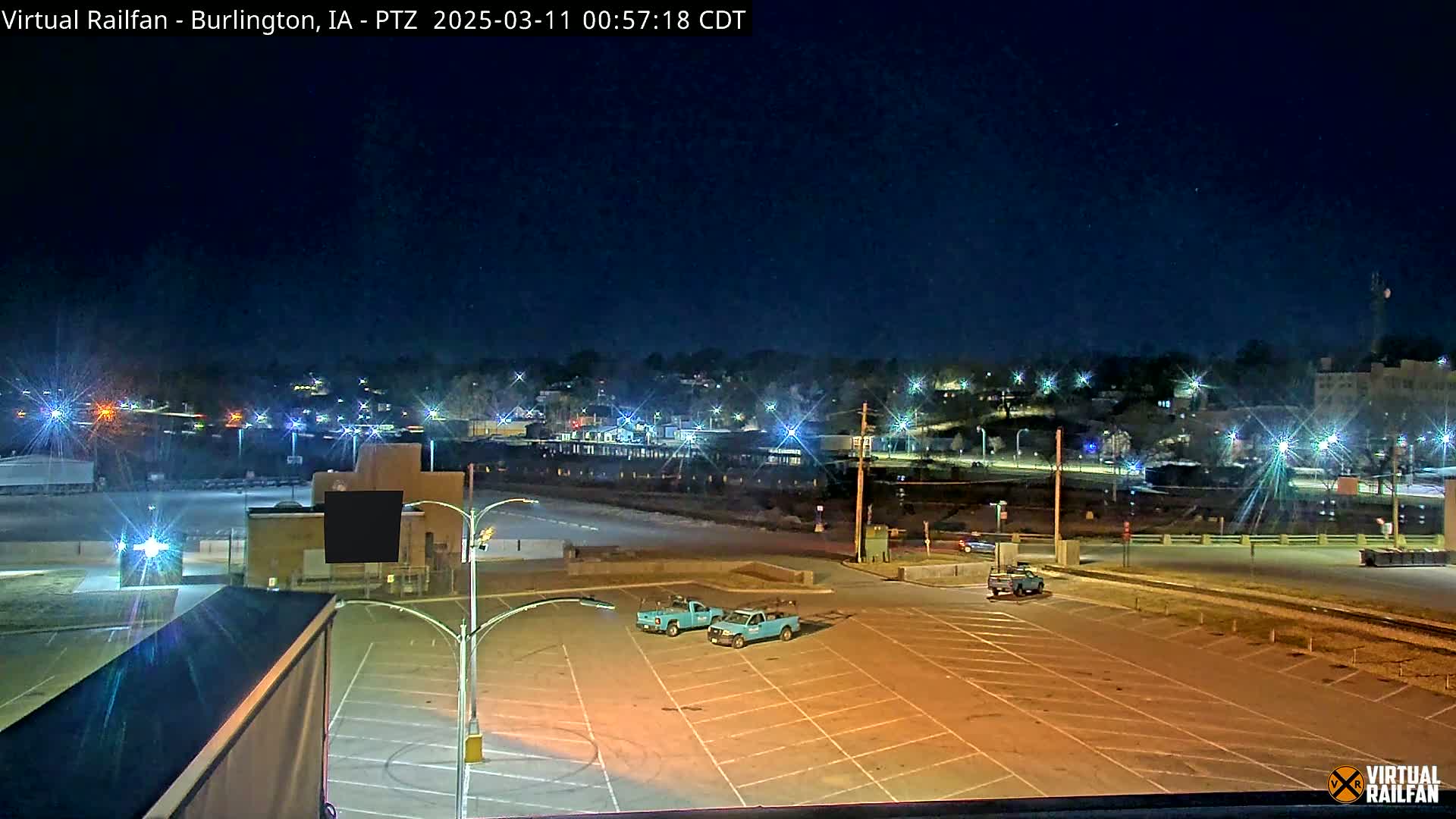 A nighttime view of a mostly empty parking lot with two light-blue trucks, overlooking a dimly lit town under a clear, dark sky.