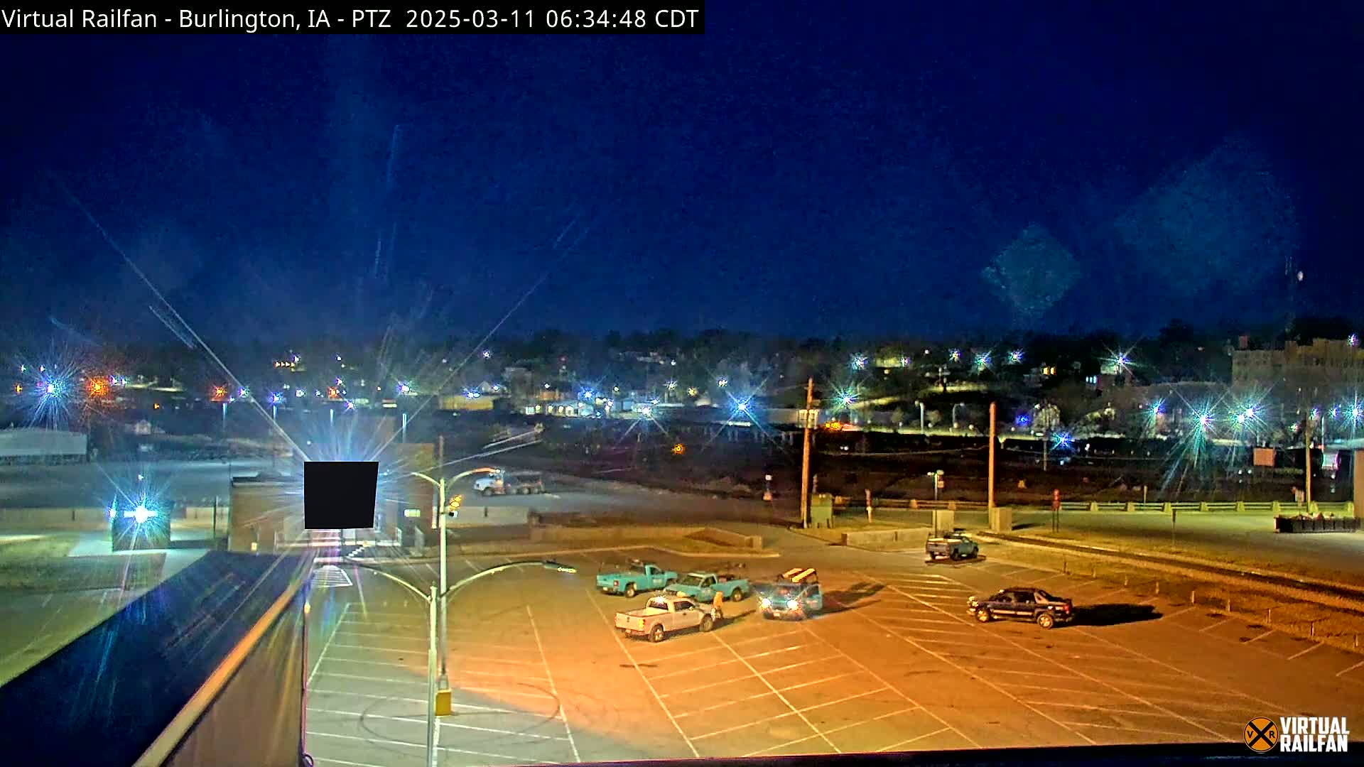 A nighttime view of an empty parking lot with several trucks and a distant town illuminated under a clear, dark blue sky.