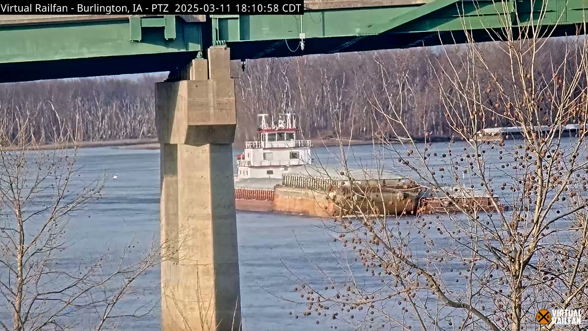 A tugboat pushing a barge travels on a river under a bridge on a sunny day, with bare trees lining the riverbank.
