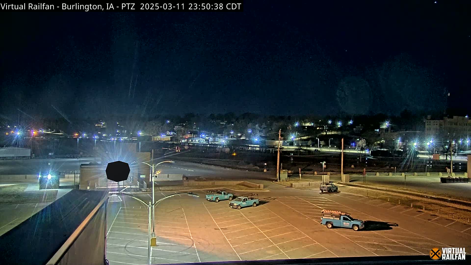 A nighttime view of an empty parking lot with several light-blue pickup trucks, overlooking a town illuminated by streetlights under a clear, starry sky.