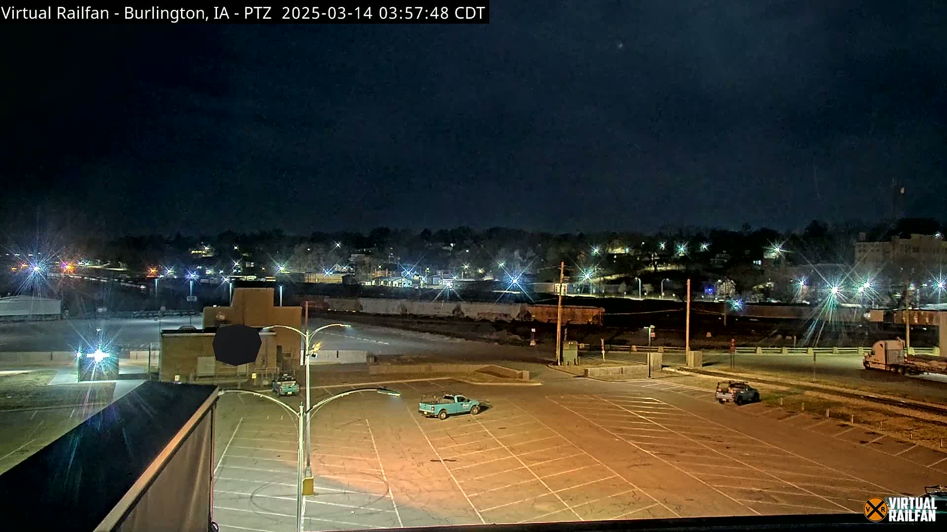 A nighttime view of an empty parking lot with a single teal truck, overlooking a railway and a dimly lit town under a dark sky.