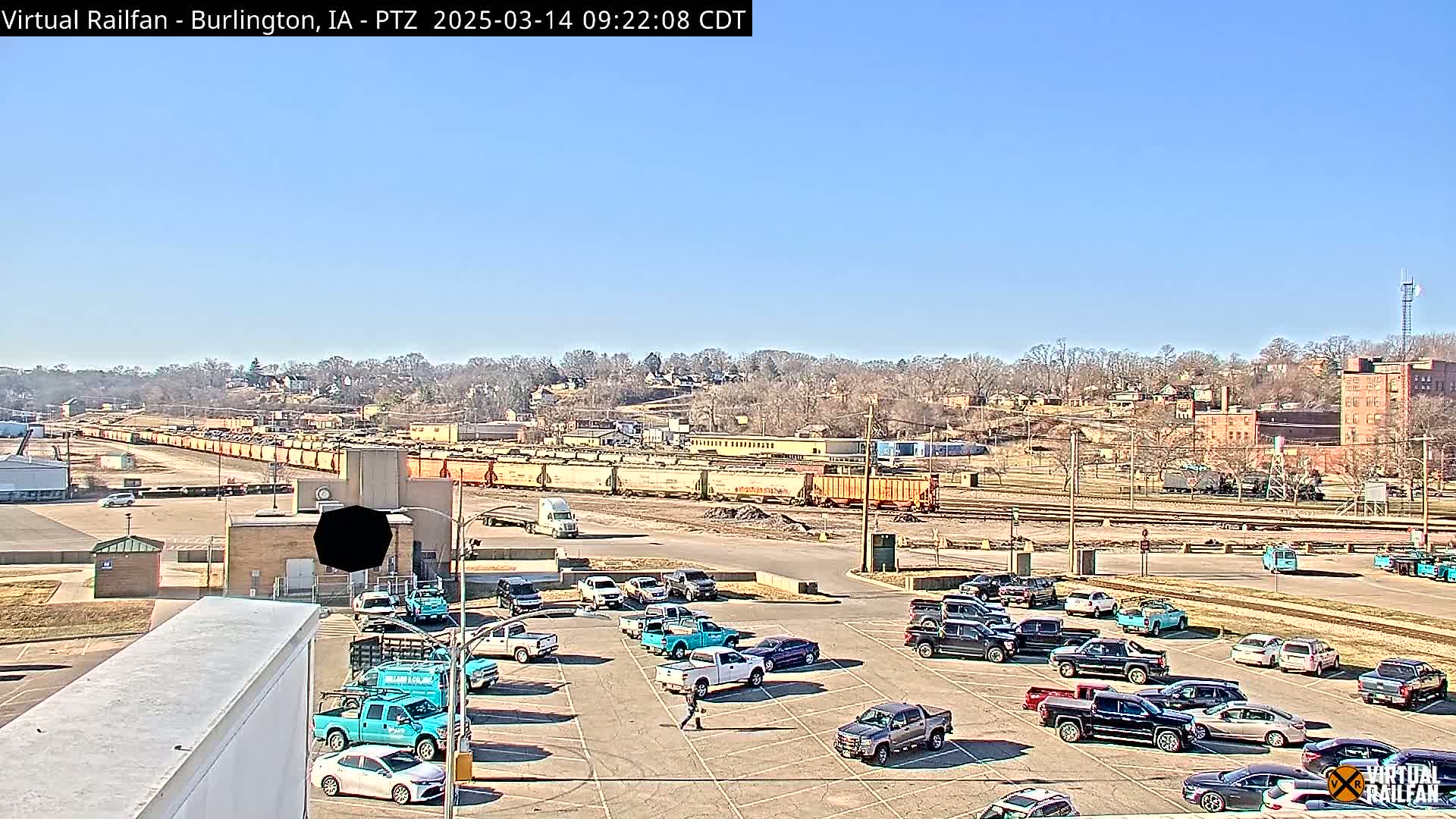 A sunny day reveals a train yard with numerous railcars, a parking lot filled with vehicles, and a residential area on a hillside in the background.