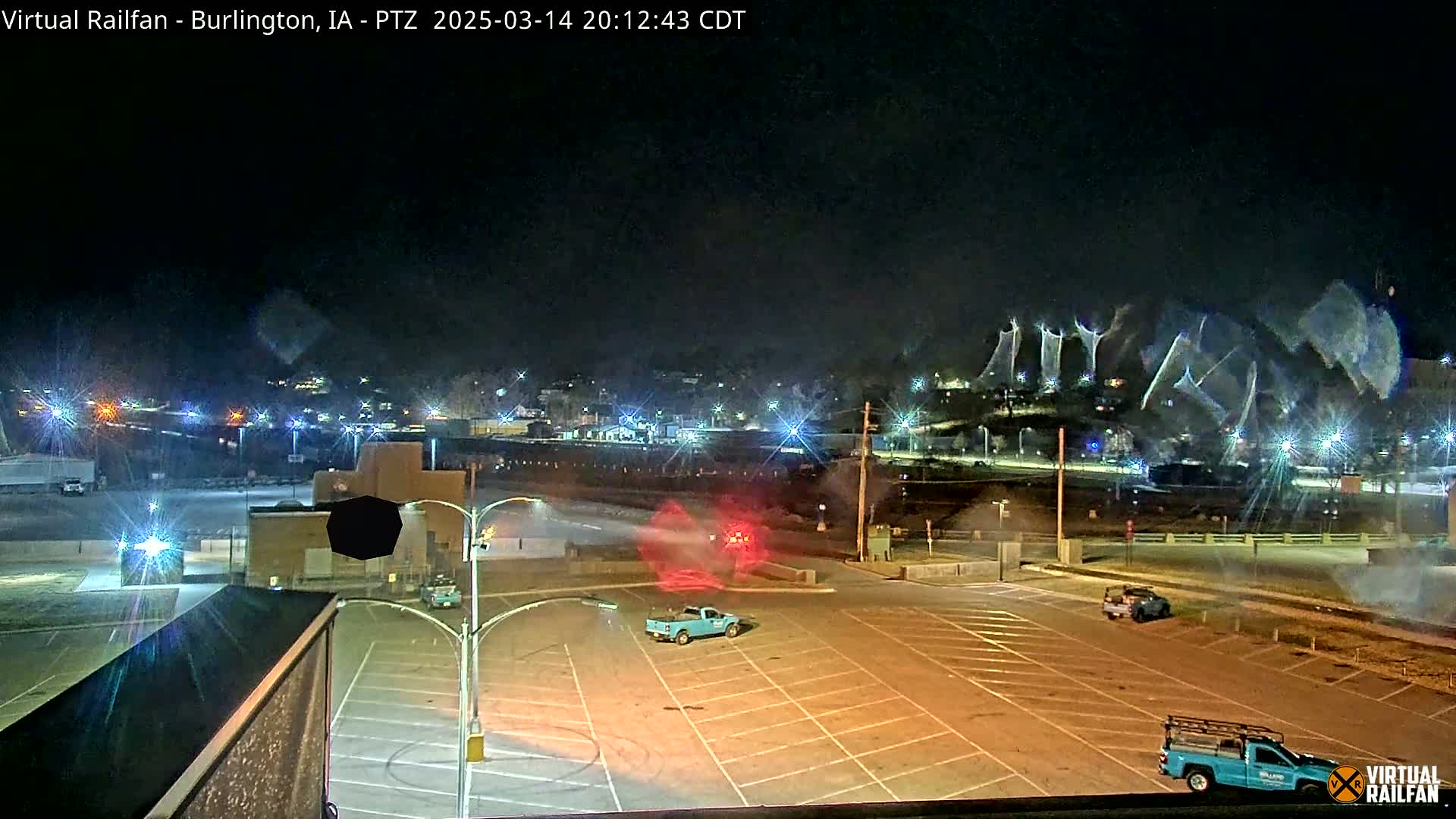 A nighttime, snow-covered parking lot with several vehicles, and a town lit up in the background under a dark, snowy sky.
