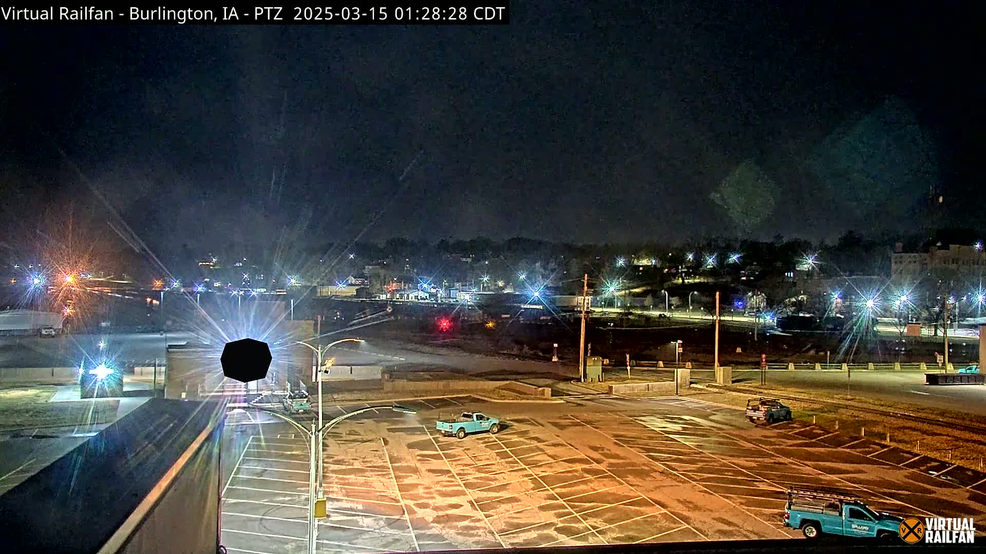 A nighttime view of a mostly empty parking lot with a few trucks, showing a town lit up in the distance under a clear, dark sky.