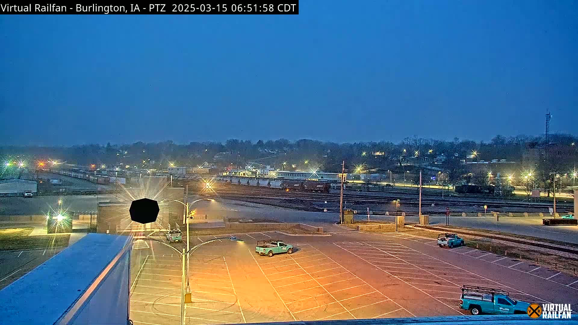 An empty parking lot at twilight, with a few trucks parked, overlooks a railroad yard and a town lit by streetlights under a clear, dark blue sky.