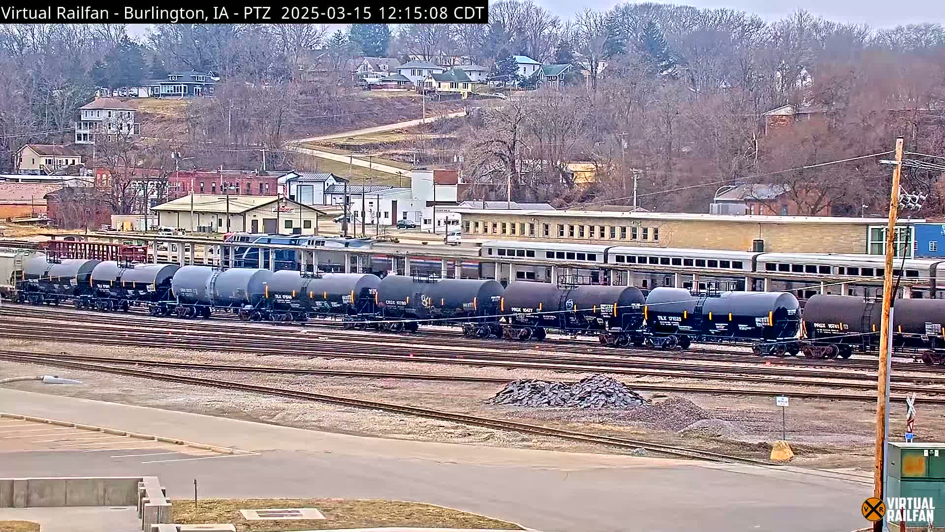A long line of dark gray tanker cars sits on a rail yard next to a town under an overcast sky.