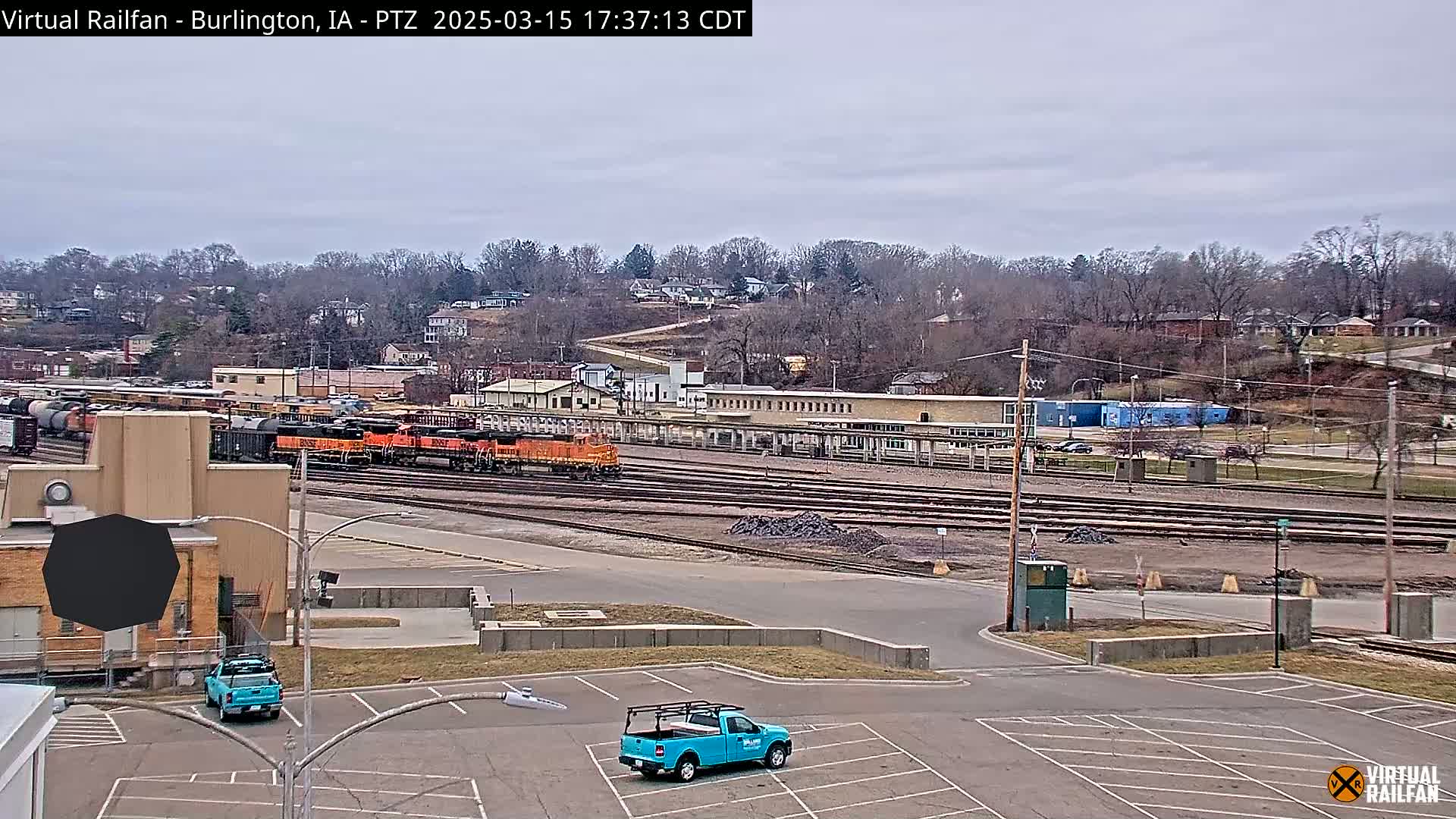 A long shot of a train yard on an overcast day, with several locomotives and freight cars visible, and a couple of light blue pickup trucks parked in an empty lot in the foreground.