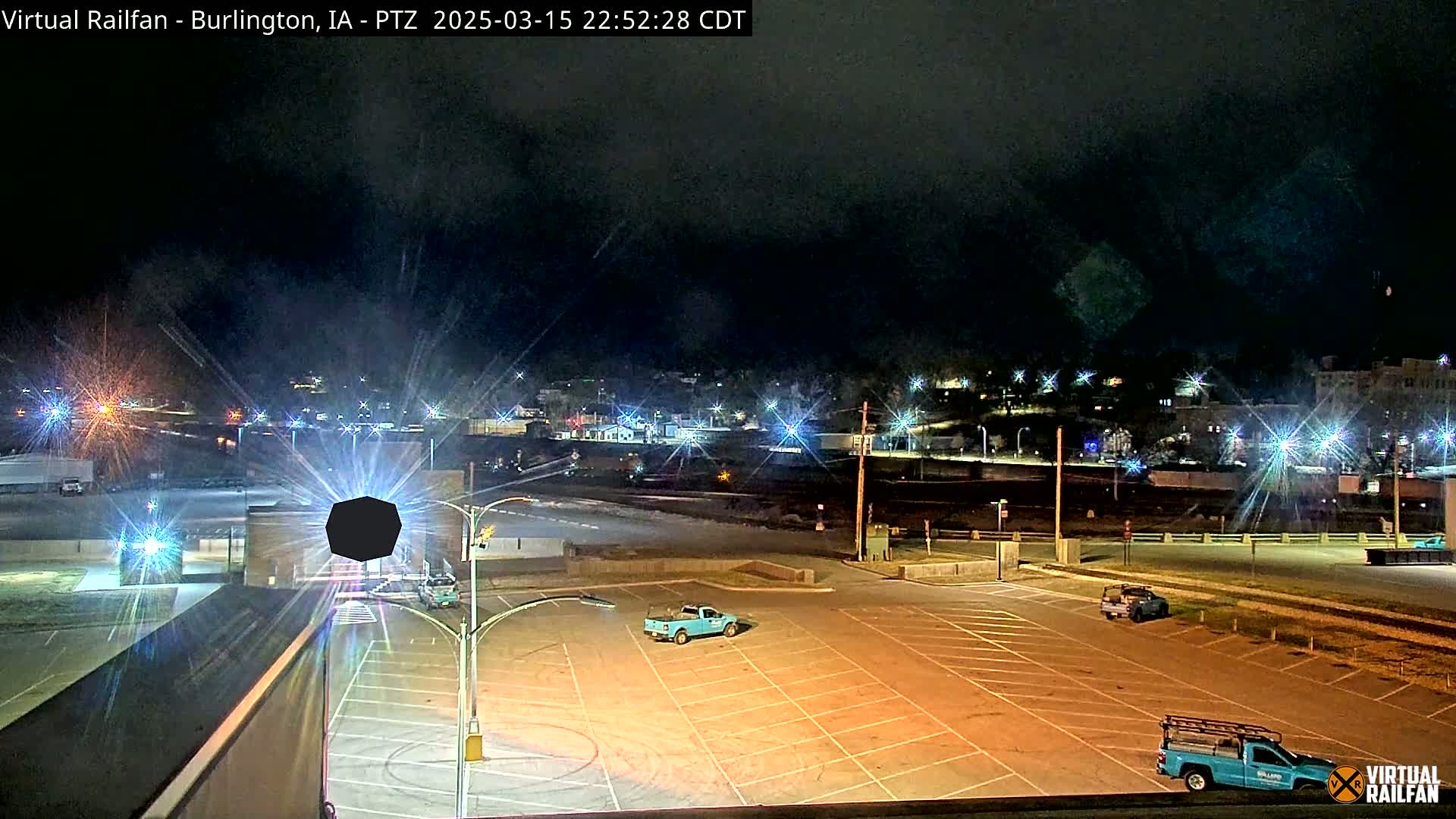 A mostly empty parking lot at night is illuminated by streetlights, with a few vehicles present and a distant town visible under a dark sky.