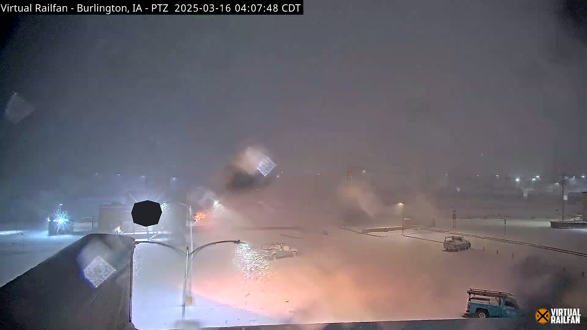 A snowstorm blankets a mostly empty roadway and parking lot at night, with a few vehicles visible amidst blowing snow.