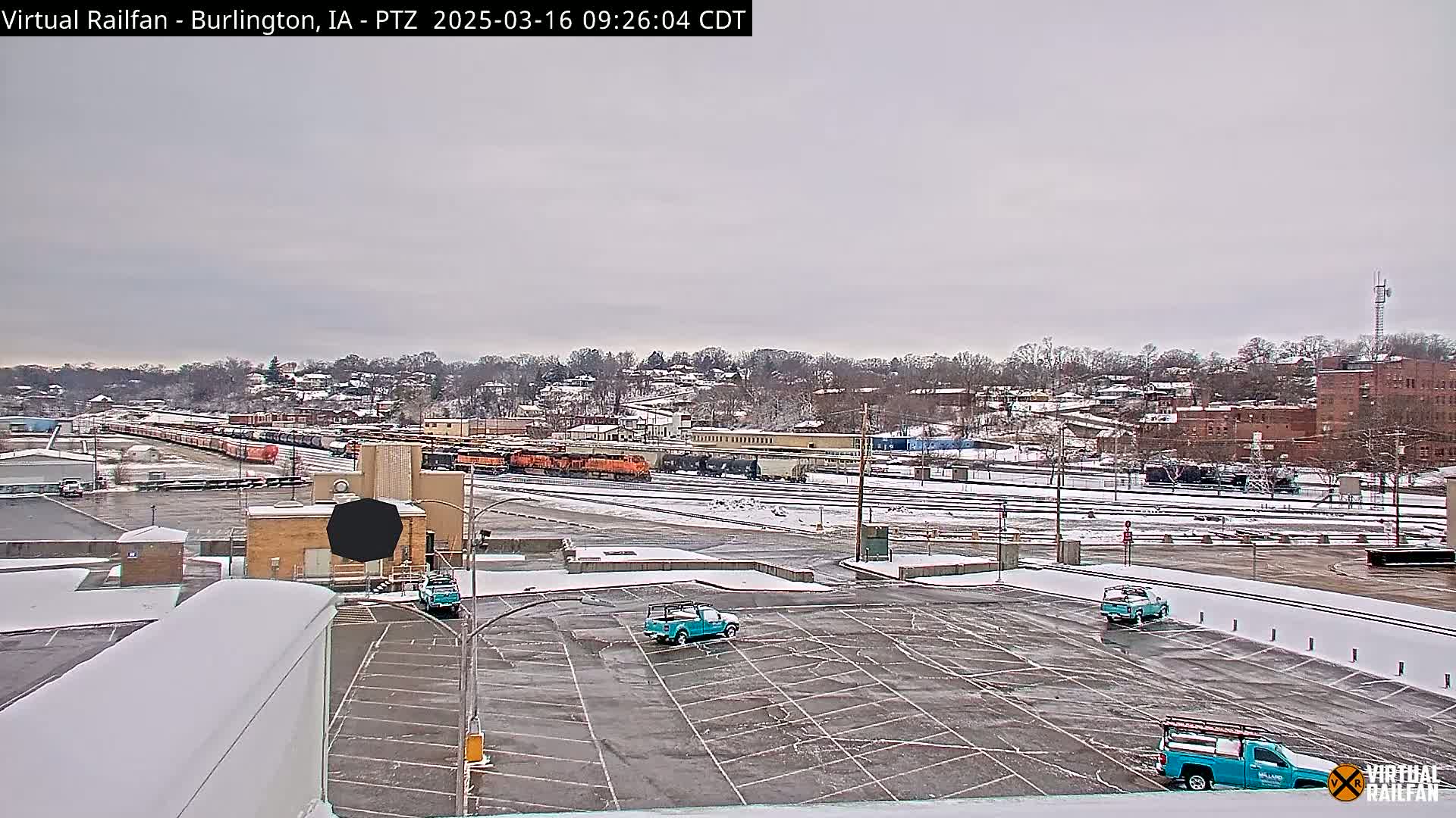 A snow-covered rail yard with several trains and a few light teal pickup trucks parked in a nearby lot, under an overcast sky.