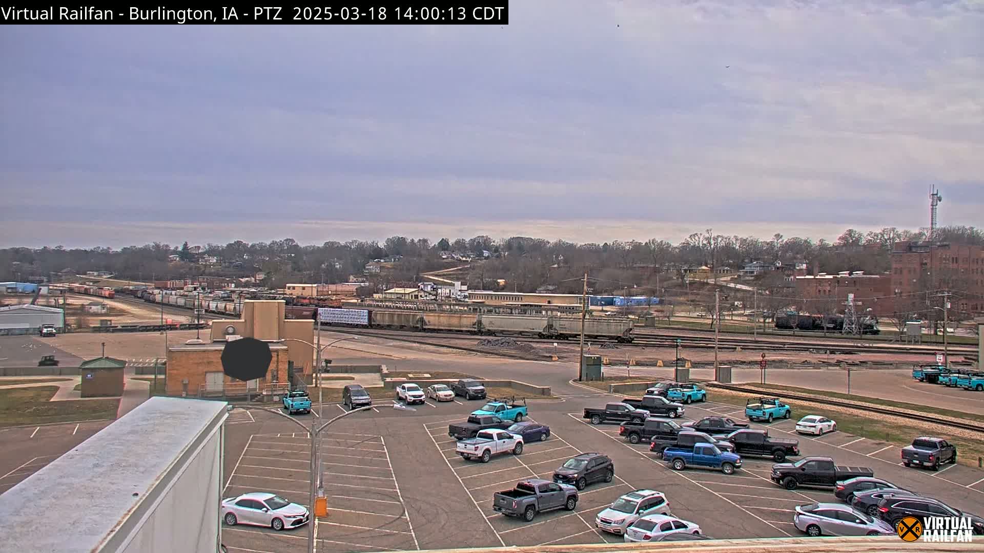 A mostly cloudy day reveals a train yard with several rail cars and a parking lot filled with numerous vehicles, all situated in front of a residential area on a hillside.