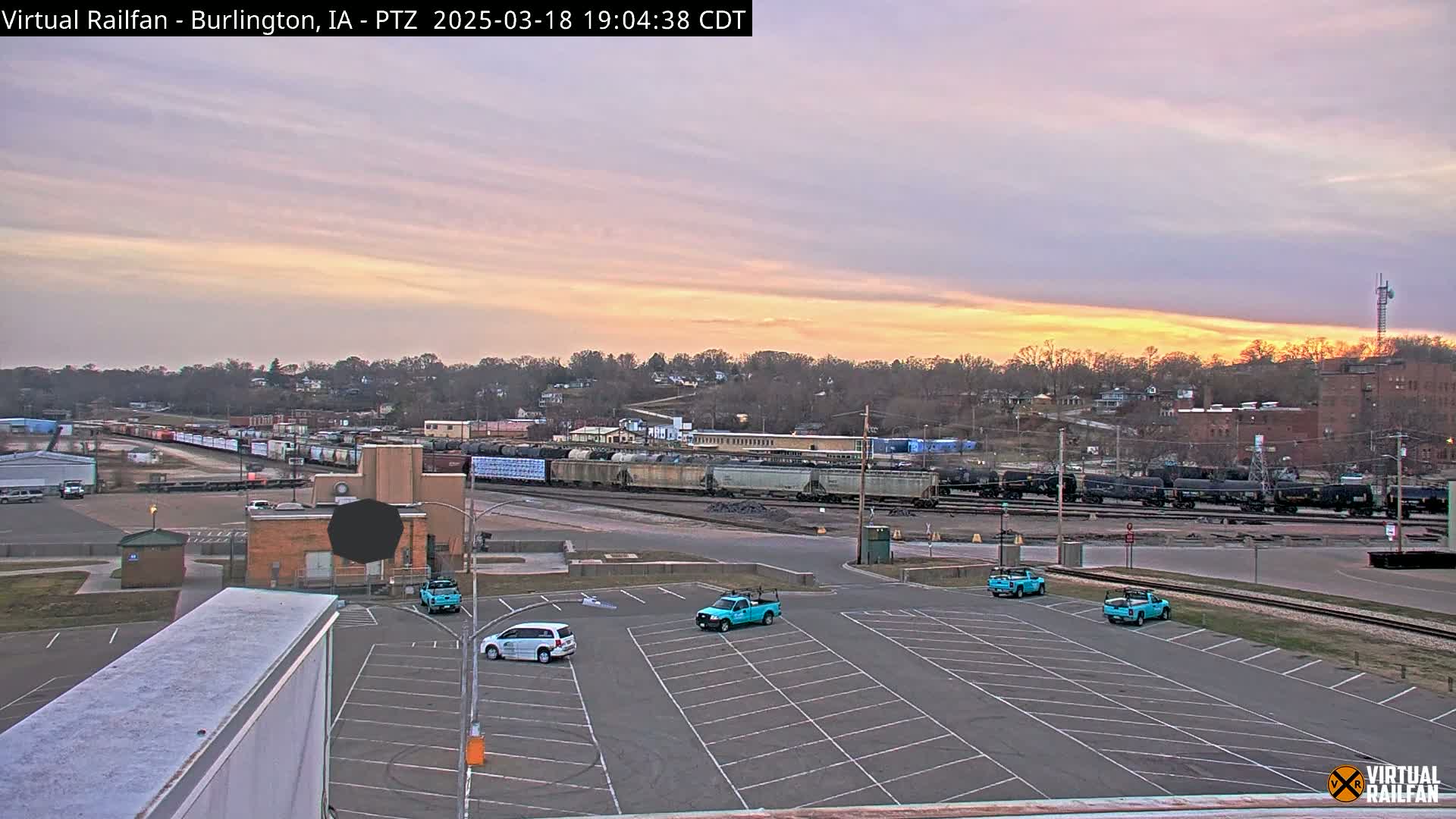 A twilight sky over a railroad yard with several parked teal trucks and cars in an empty parking lot.
