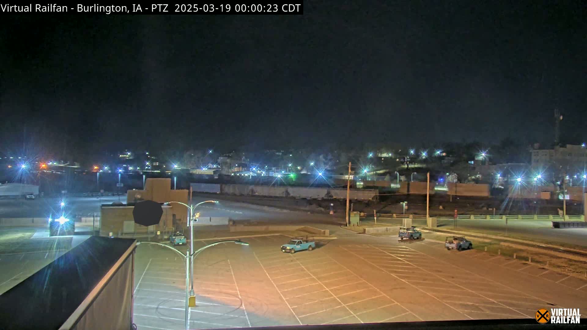 A nighttime view of a mostly empty parking lot, with a few vehicles present, next to a rail yard and a dimly lit town in the background under a clear sky.