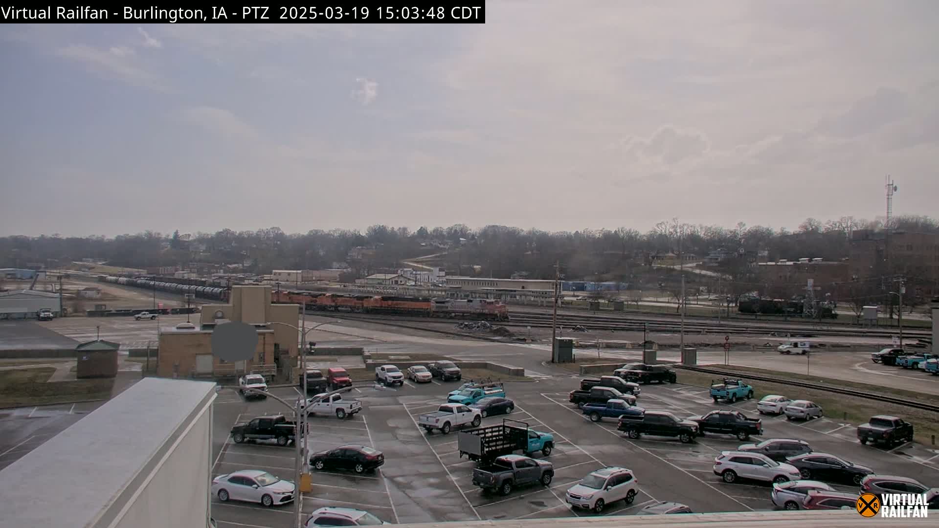 A mostly cloudy day reveals a train yard with several locomotives and rail cars, a parking lot full of vehicles, and a treeline in the background.