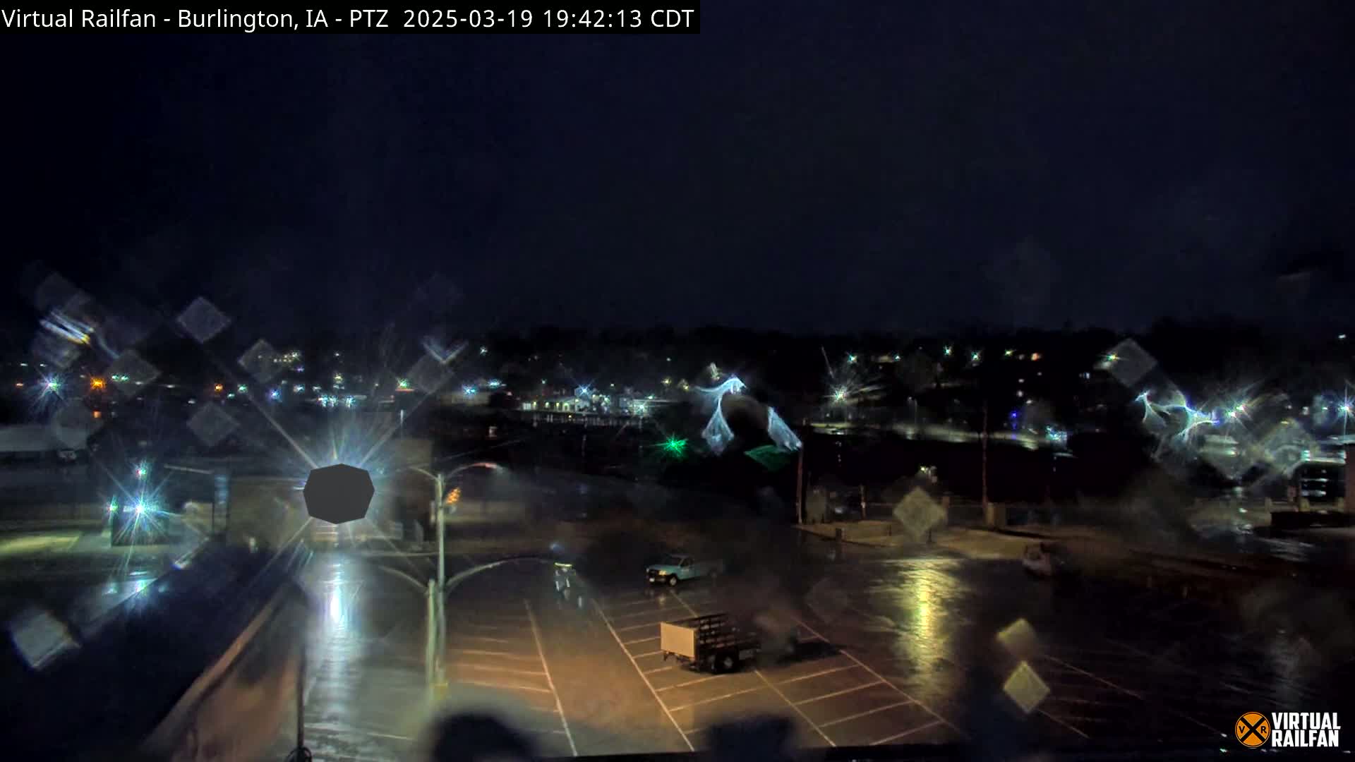 A nighttime view of a rain-slicked parking lot and street, with a few vehicles visible, under a dark, overcast sky.