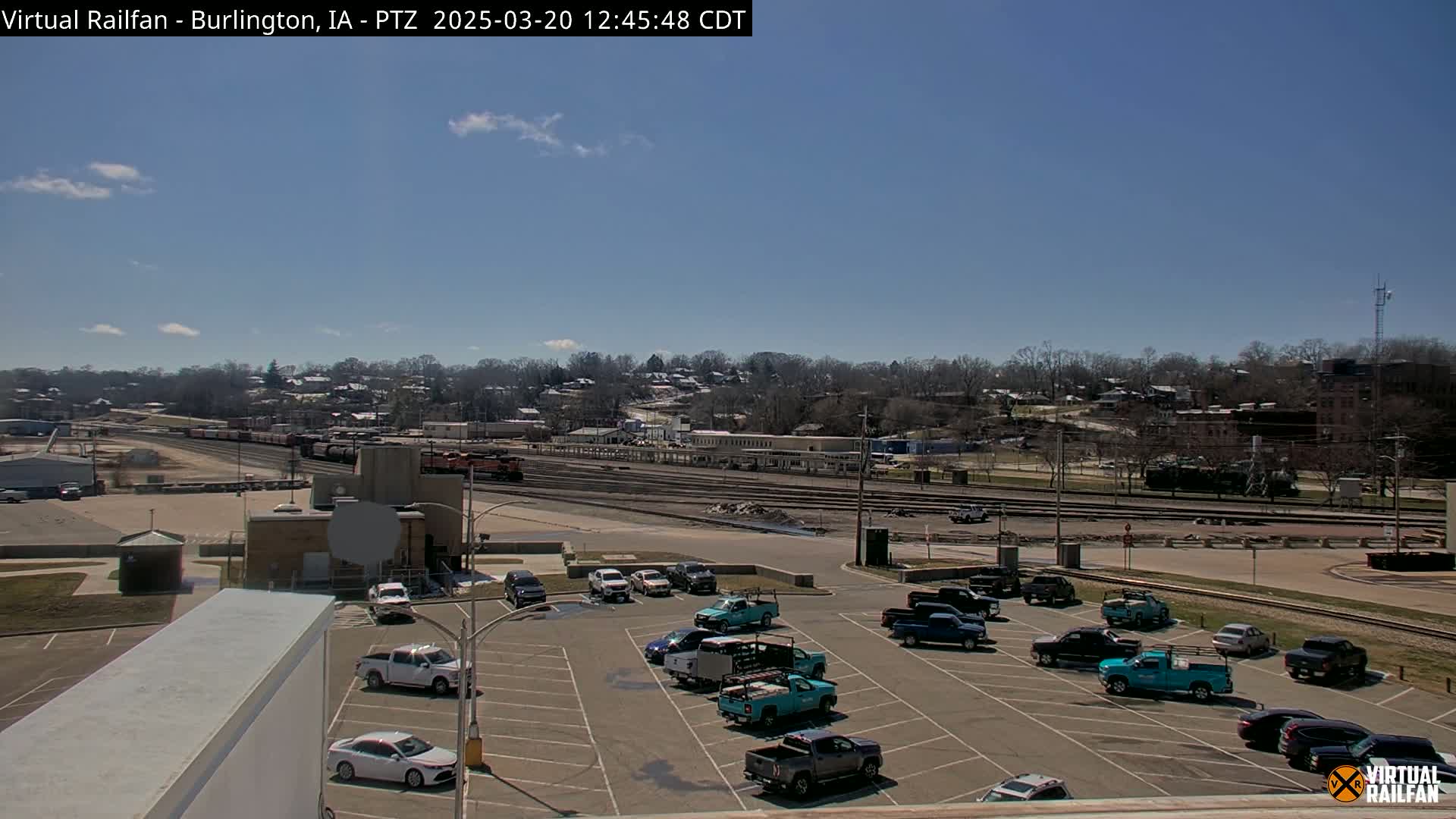 A mostly sunny day shows a large parking lot filled with pickup trucks and cars, overlooking a train yard and a residential area beyond.