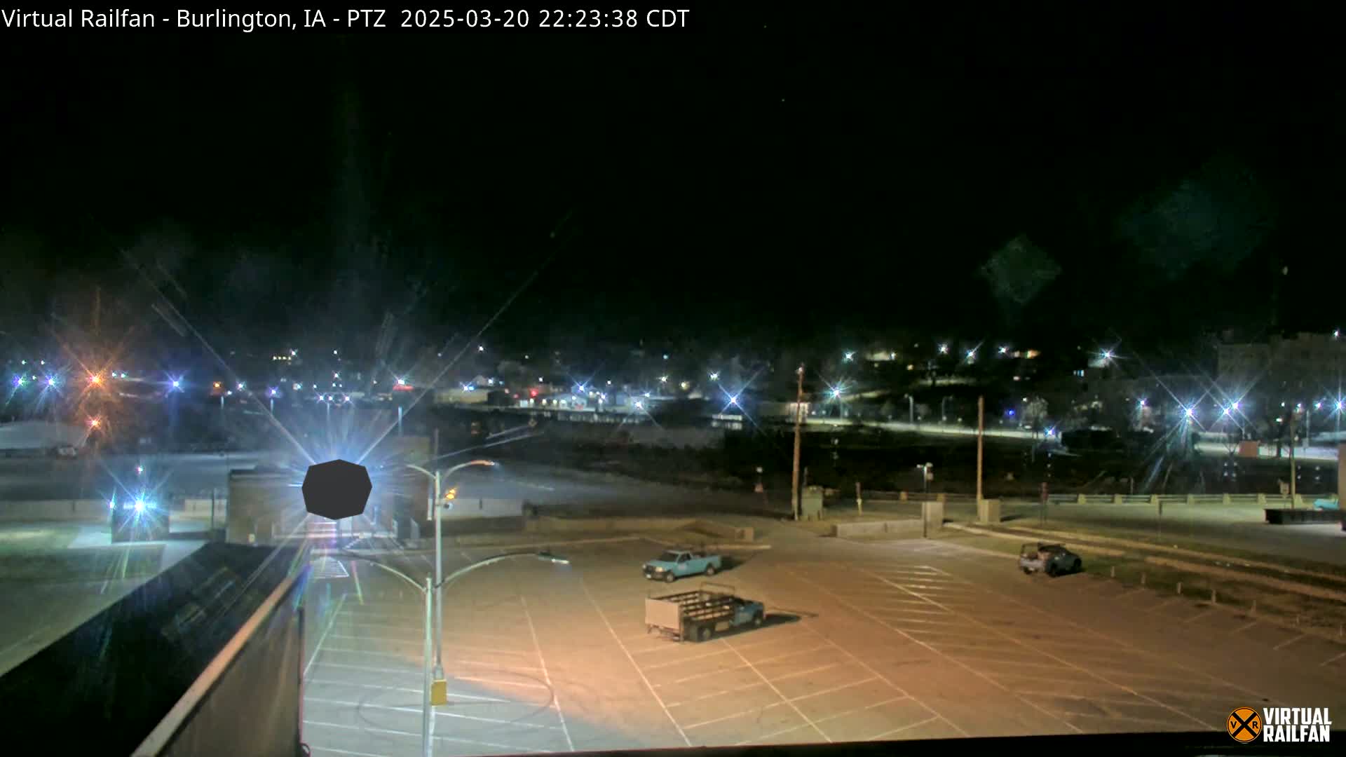 A nighttime view of an empty parking lot with a few vehicles, illuminated by streetlights, overlooks a distant town also lit up at night.