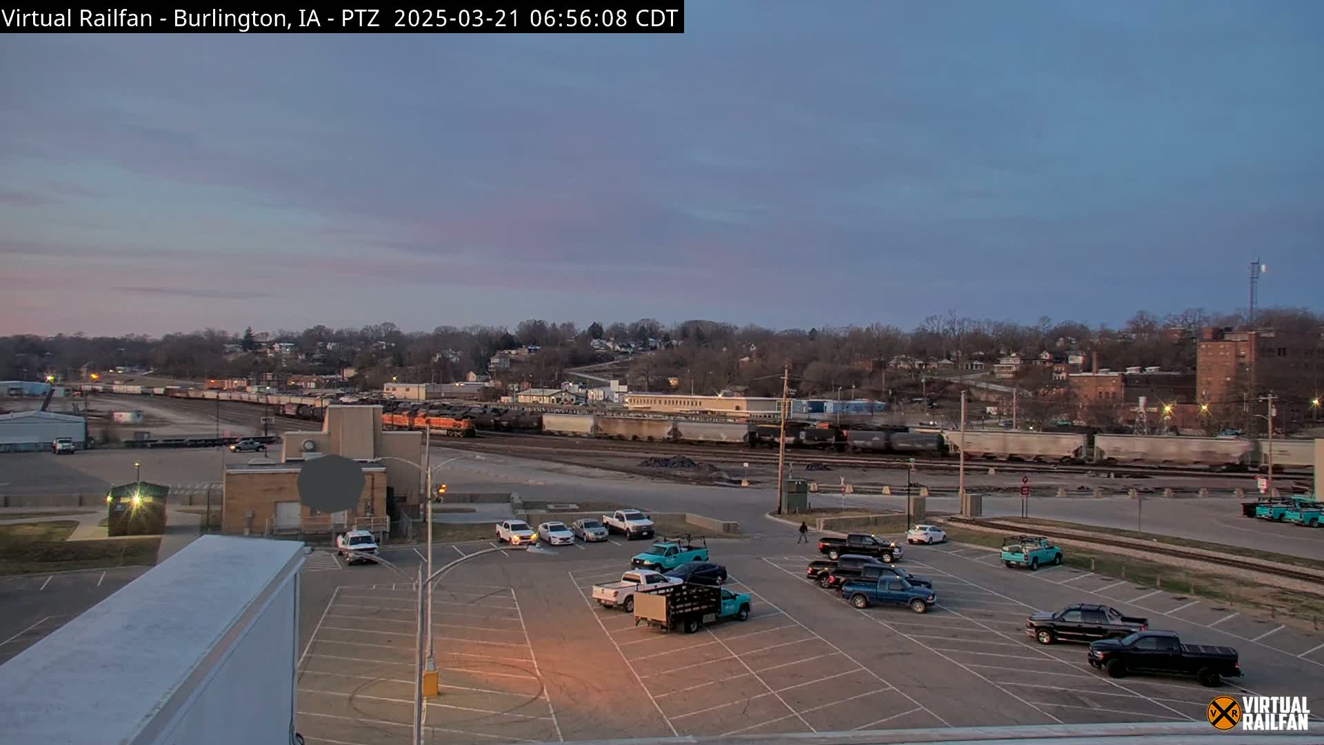 A twilight view of a railroad yard with numerous parked cars and trucks in an adjacent parking lot, under a clear, dusky sky.