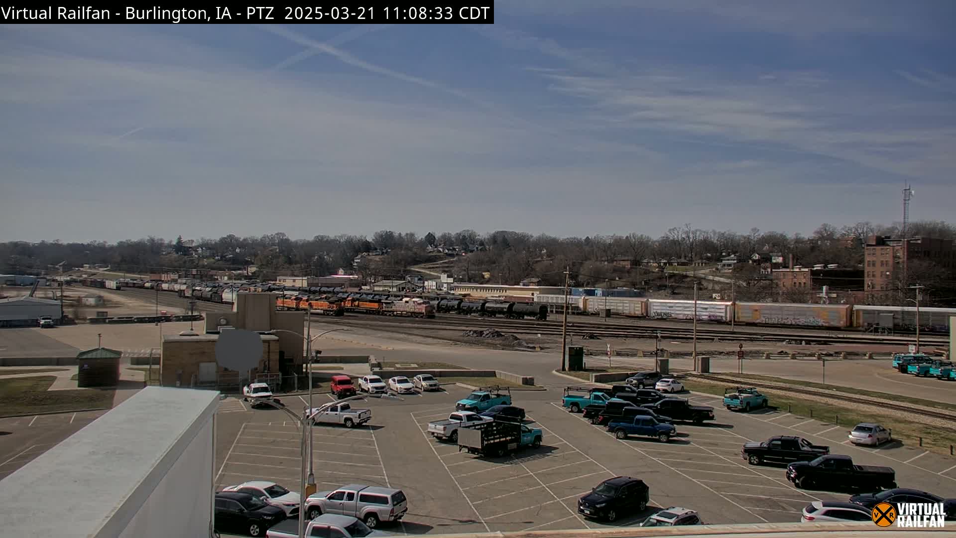 A long freight train is parked near a parking lot filled with various trucks and cars under a mostly sunny sky.