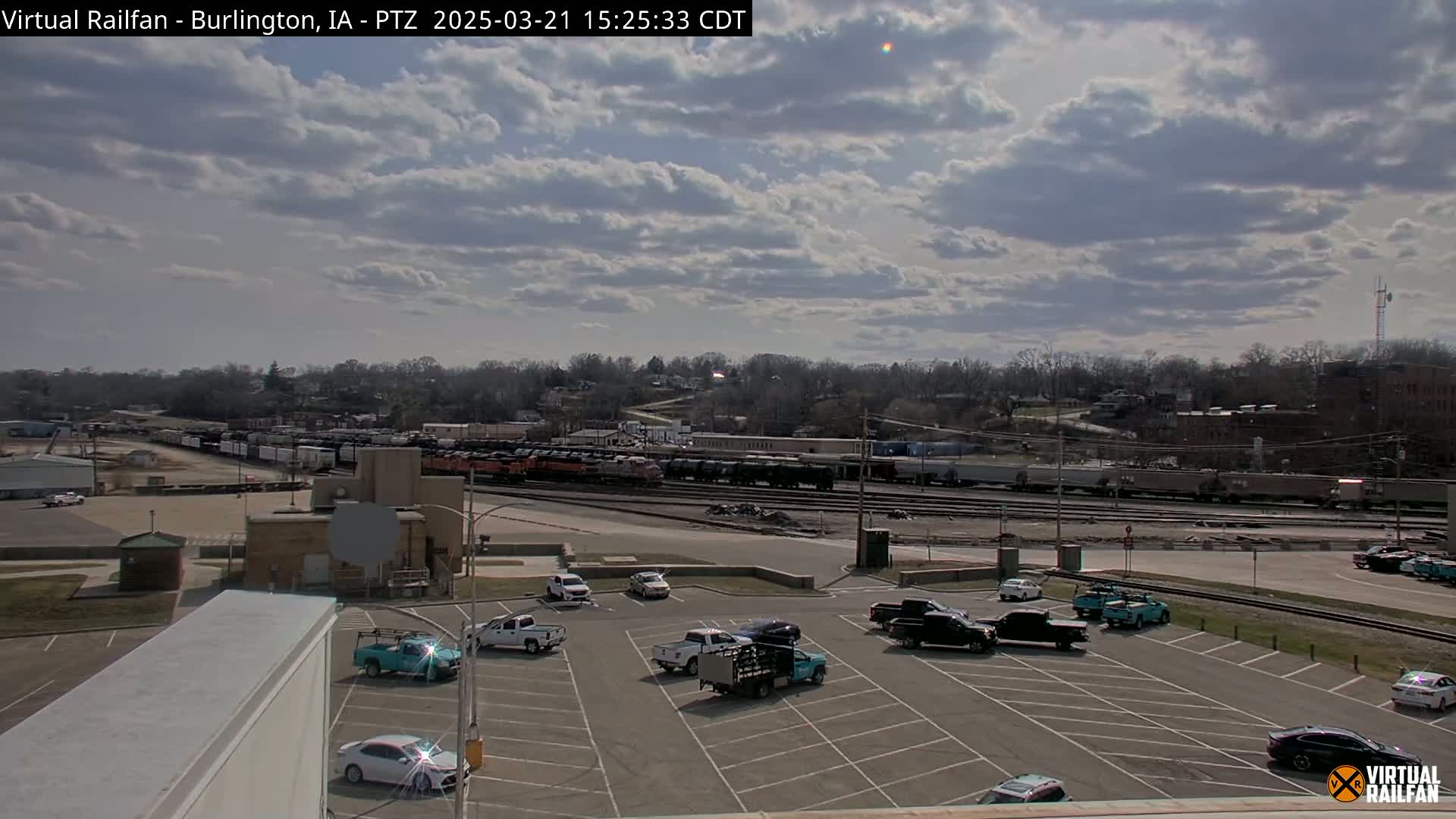 A partly cloudy sky overlooks a train yard with numerous railcars and a parking lot containing several pickup trucks and cars.