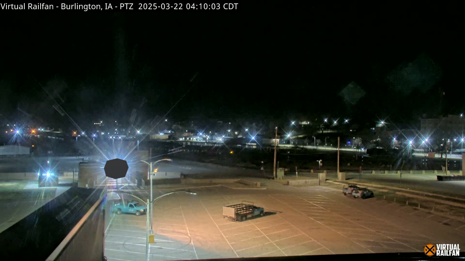 A nighttime view of an empty parking lot with a few vehicles, illuminated by streetlights, overlooking a distant town also lit up under a clear, dark sky.