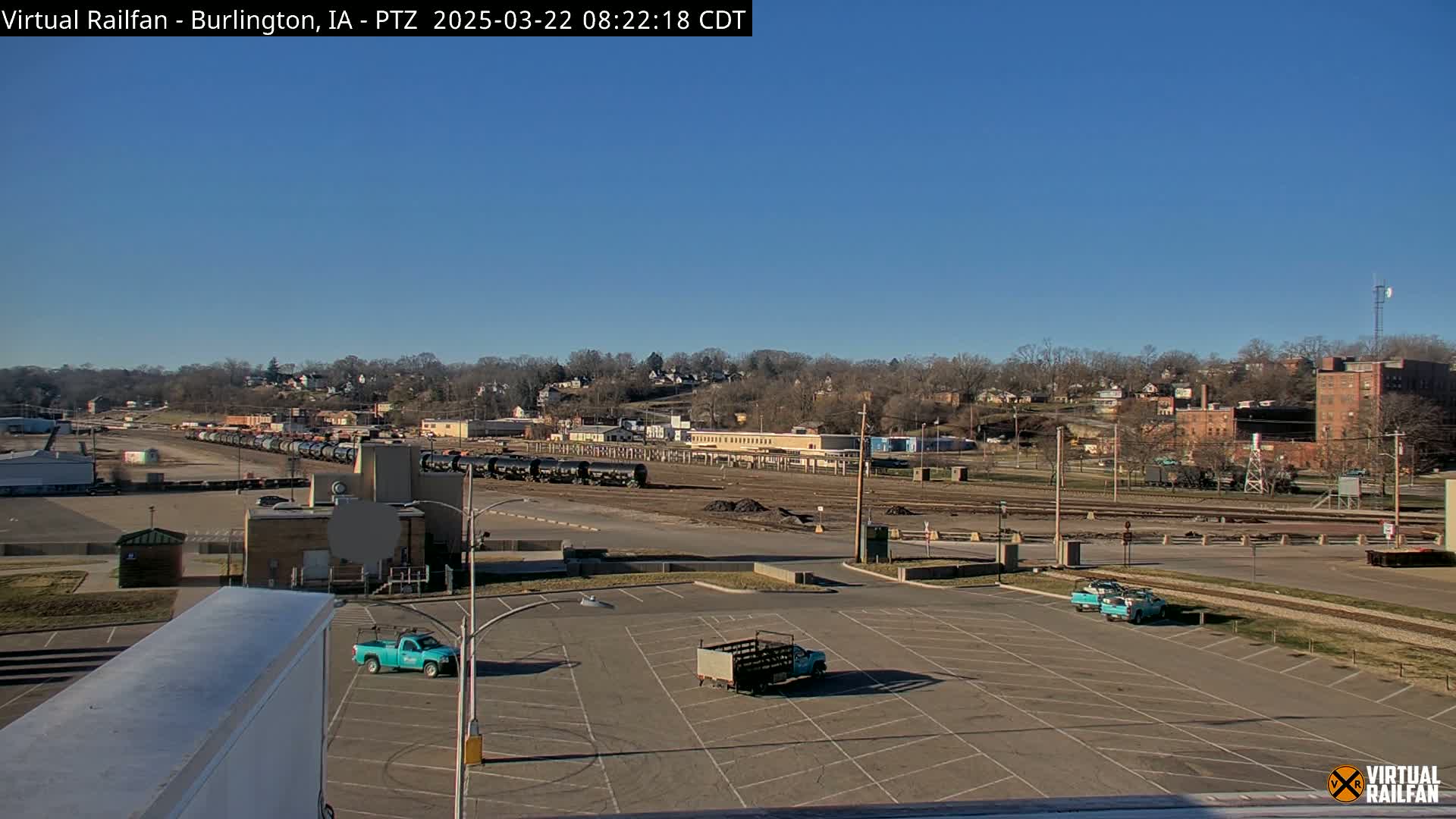 A mostly empty parking lot sits in the foreground, with a train and a residential area visible in the background under a clear, sunny sky.
