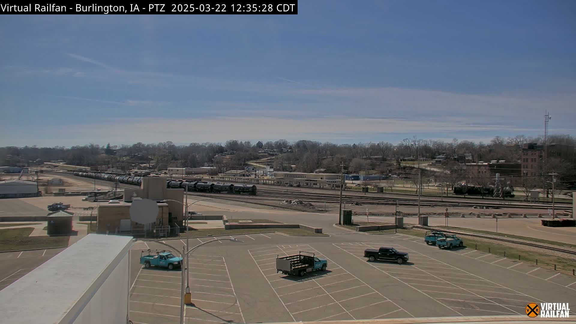 A long freight train sits in a rail yard under a clear, sunny sky, with several pickup trucks parked in an adjacent empty parking lot.