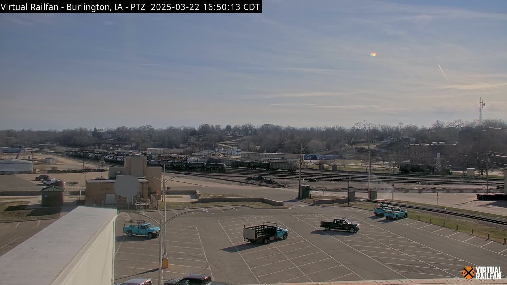 A mostly sunny day reveals a train yard with several railcars, a parking lot with pickup trucks, and a tree-lined hill in the background.