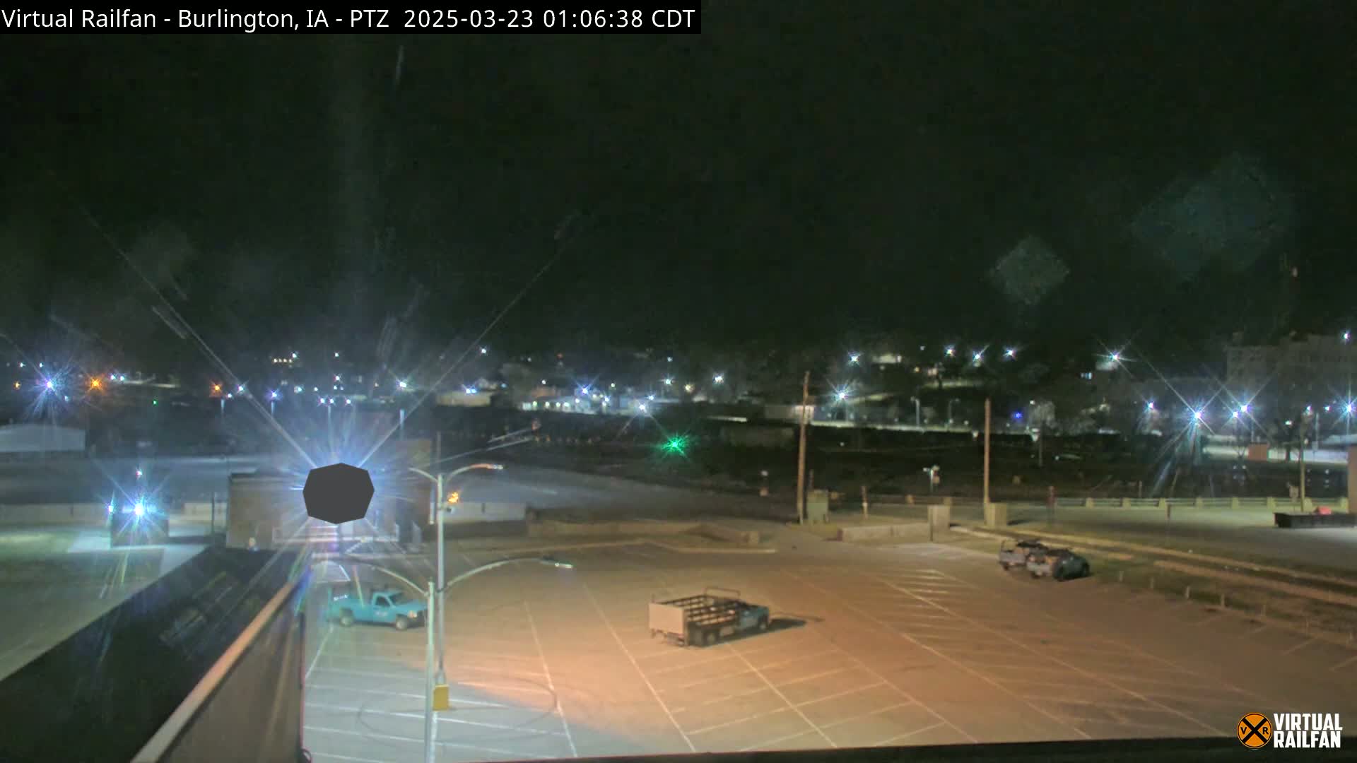 A nighttime, dimly lit view of a mostly empty parking lot with a few vehicles, overlooking a distant town with streetlights.