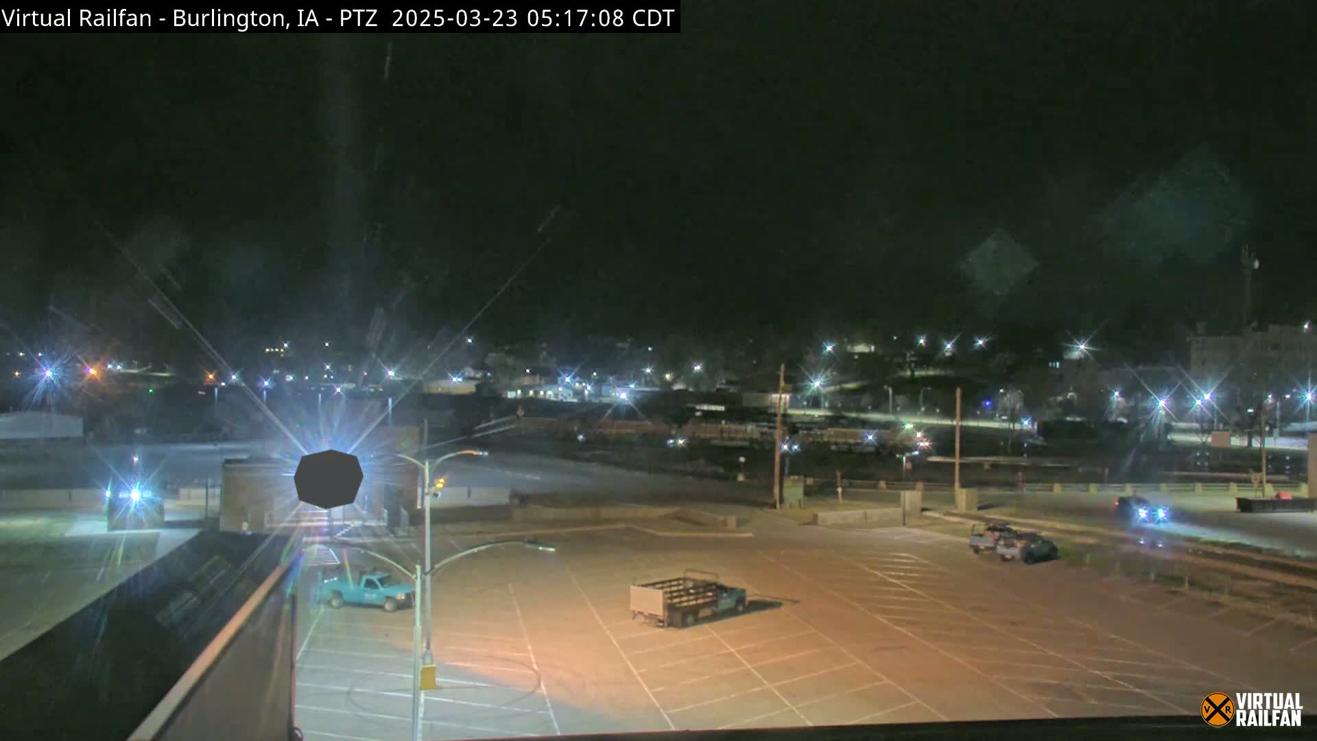 A nighttime view of a mostly empty parking lot with a few vehicles, and a distant town illuminated by streetlights under a clear, dark sky.