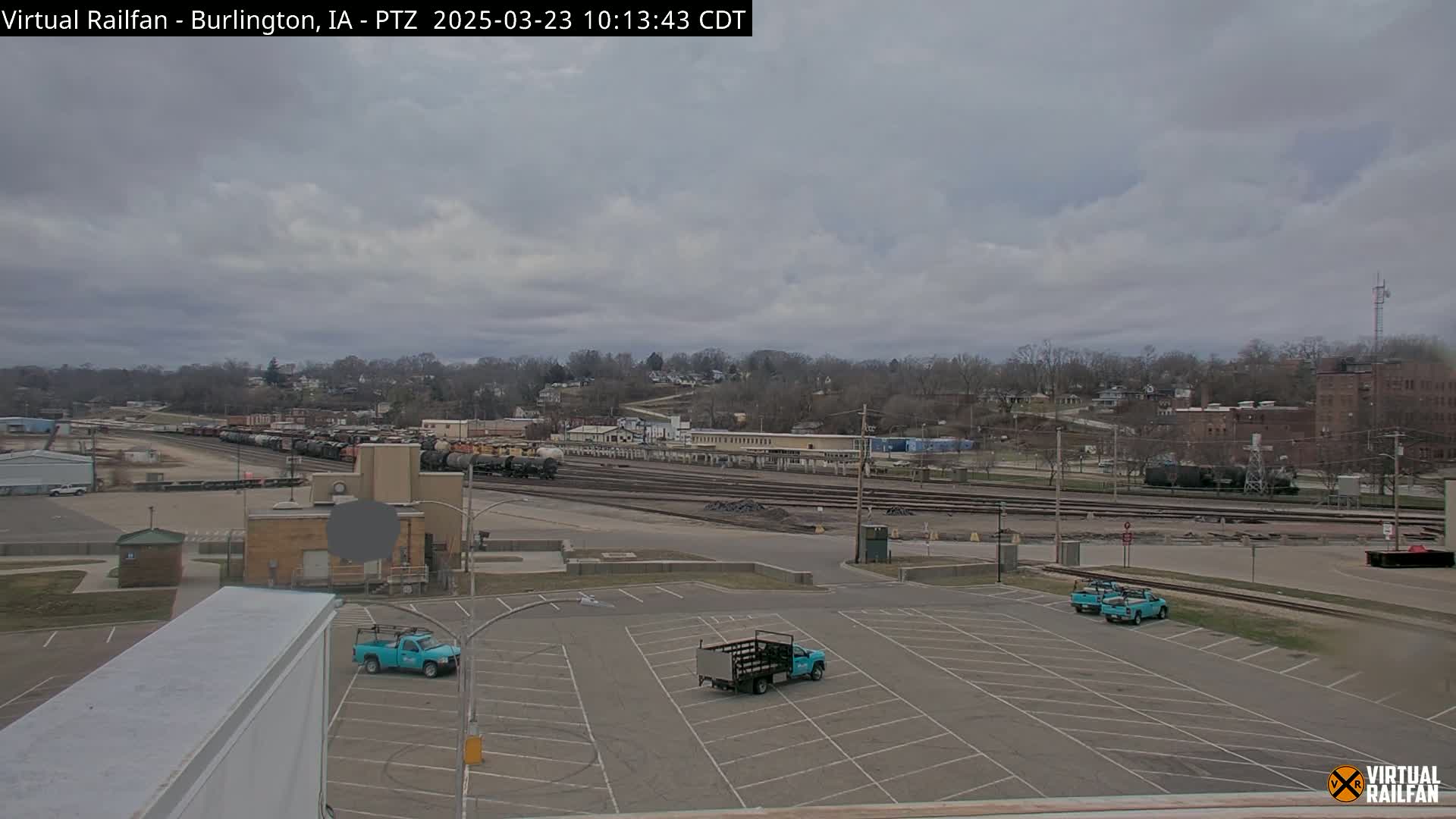 A mostly cloudy day overlooks a train yard with several rail cars and a few light-teal pickup trucks parked in an empty lot.