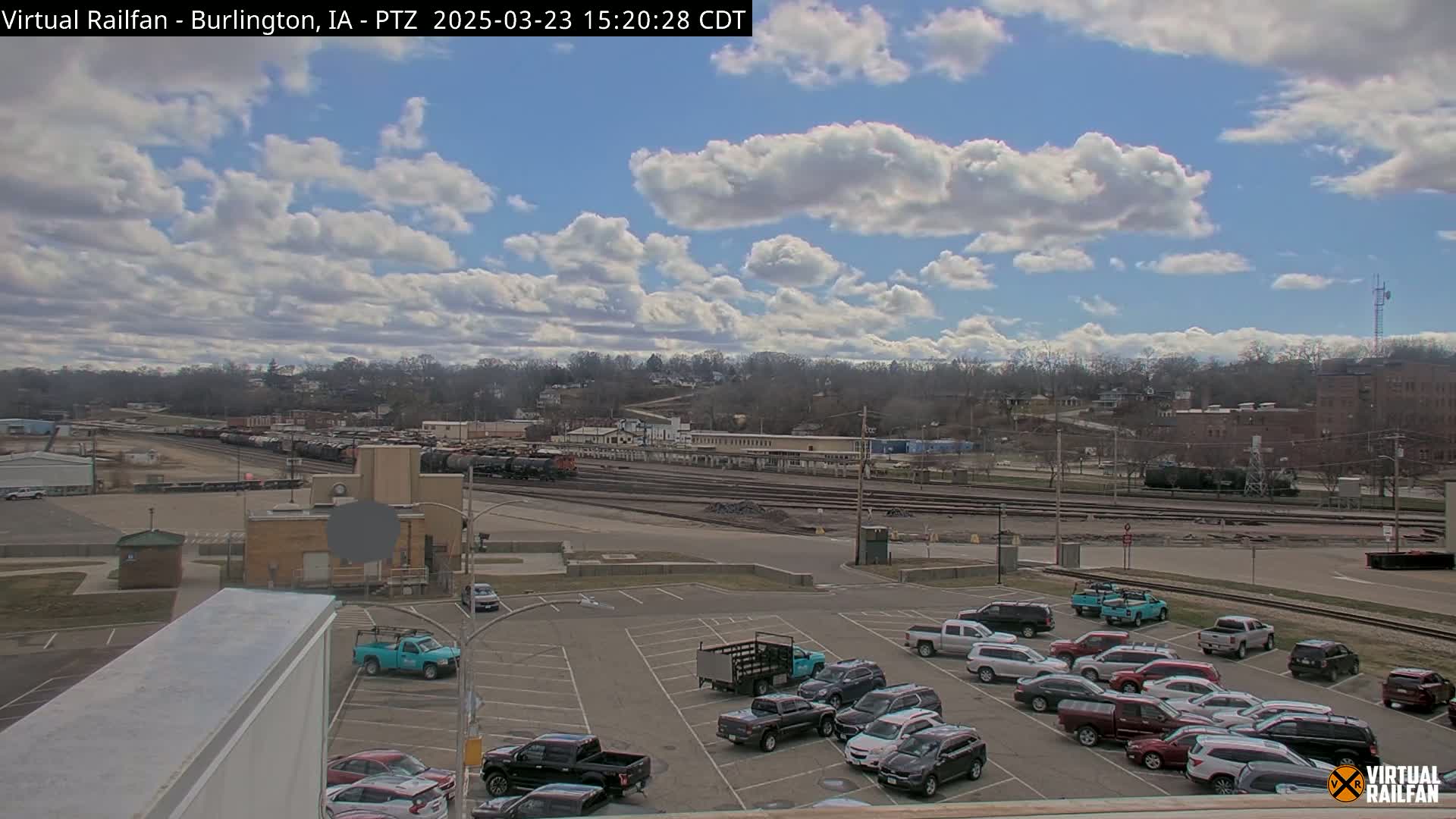 A partly cloudy day reveals a railroad yard with numerous freight cars, a parking lot filled with vehicles, and a tree-lined hillside in the background.