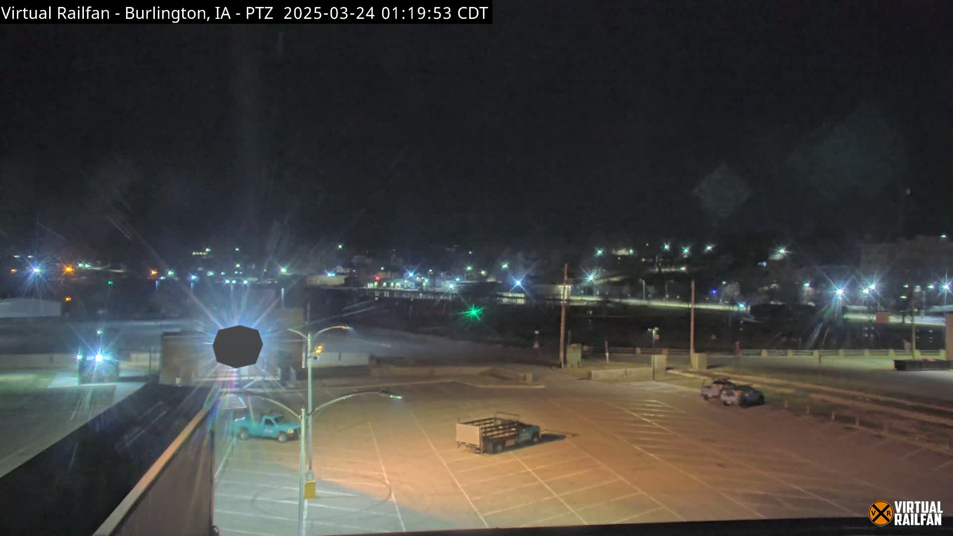 A nighttime view of a mostly empty parking lot with a few vehicles, illuminated by streetlights, overlooks a dimly lit town under a clear, dark sky.