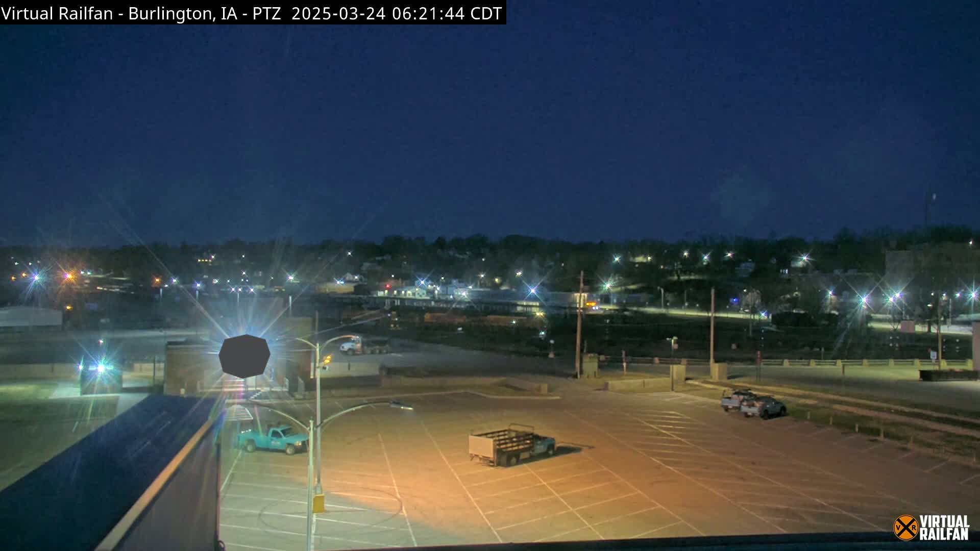 An empty, illuminated parking lot overlooks a nighttime town with a dark sky.