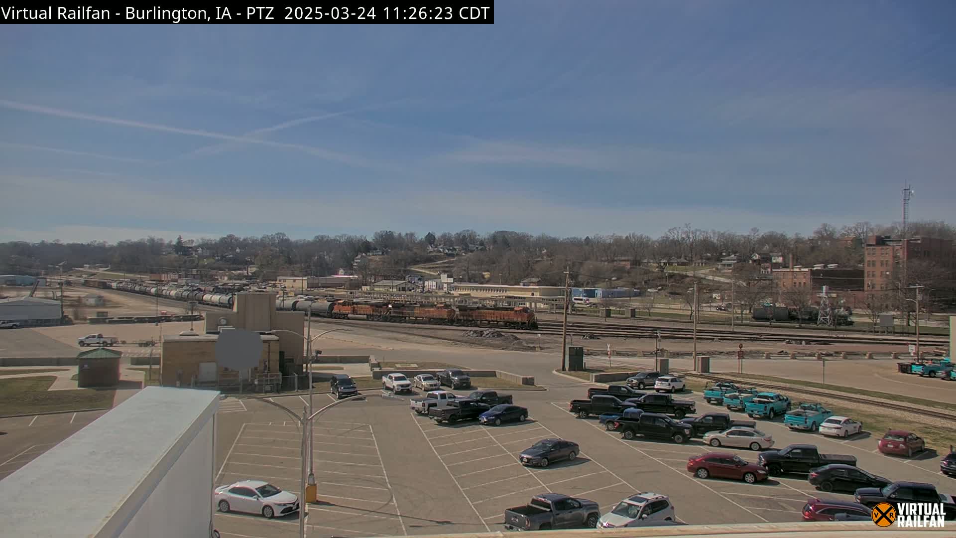 A long freight train sits in a rail yard next to a parking lot full of cars under a clear blue sky.
