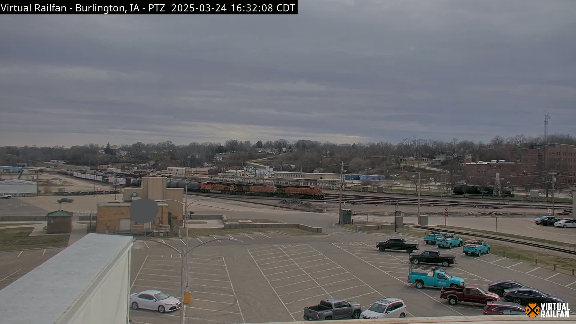 A train sits in a rail yard next to a parking lot with several pickup trucks and cars under an overcast sky.