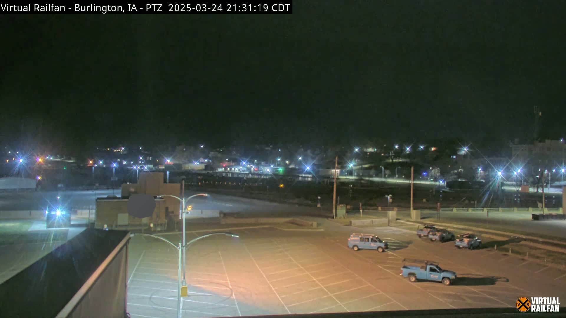 A nighttime view of an empty parking lot with several service trucks parked, and a brightly lit town in the background under a clear, dark sky.