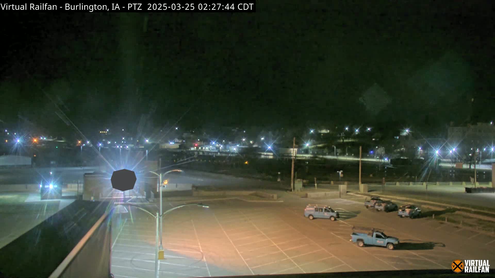 A nighttime view of an empty parking lot with several parked trucks, and a distant town illuminated by streetlights under a clear, dark sky.