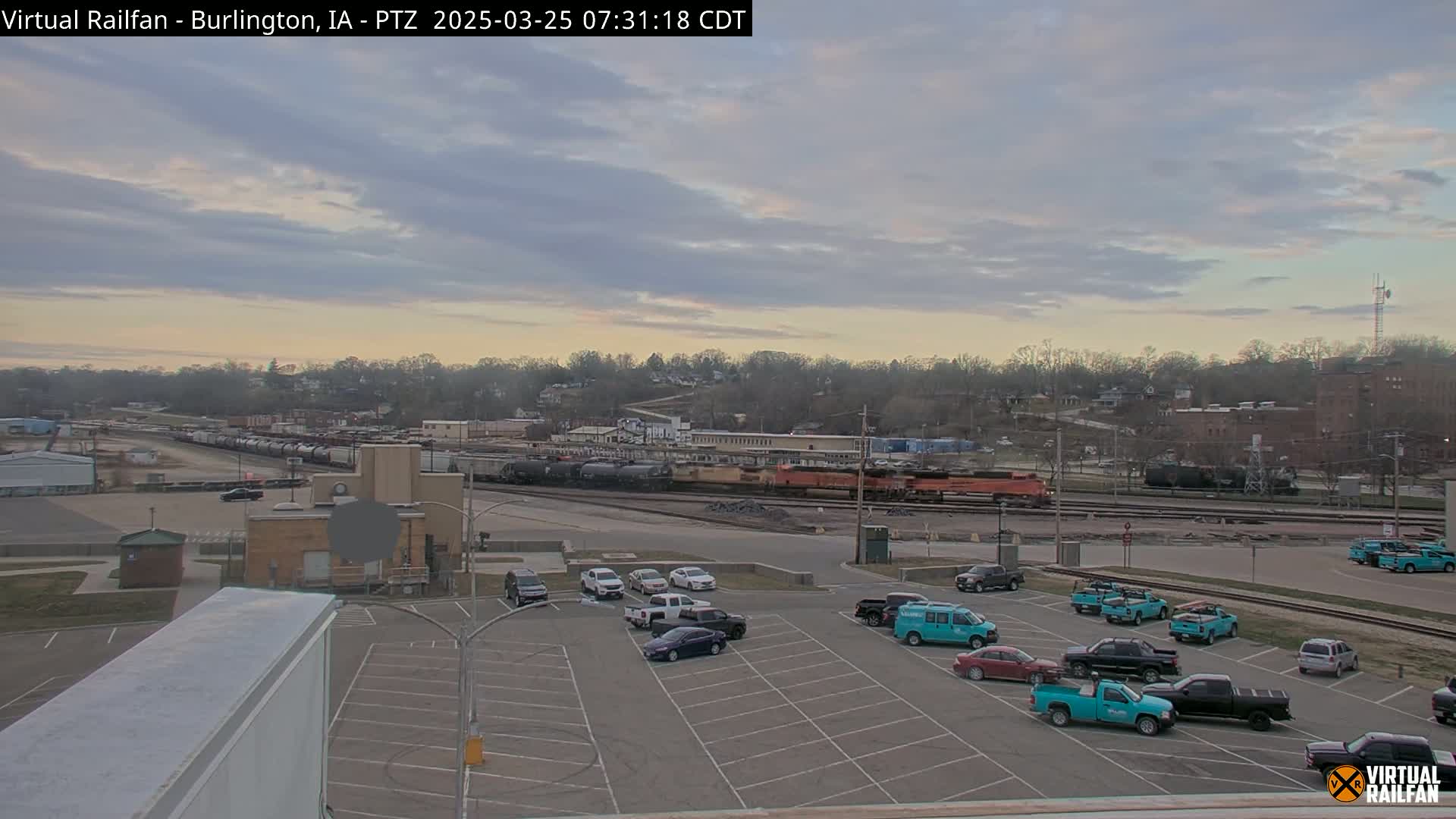 A mostly cloudy sunrise illuminates a railway yard with numerous train cars and a parking lot containing several vehicles.