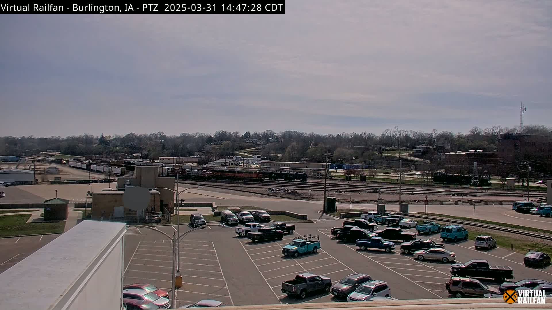 A mostly sunny day shows a railway yard with several train cars and a parking lot filled with numerous vehicles, all situated beside a tree-lined hillside.