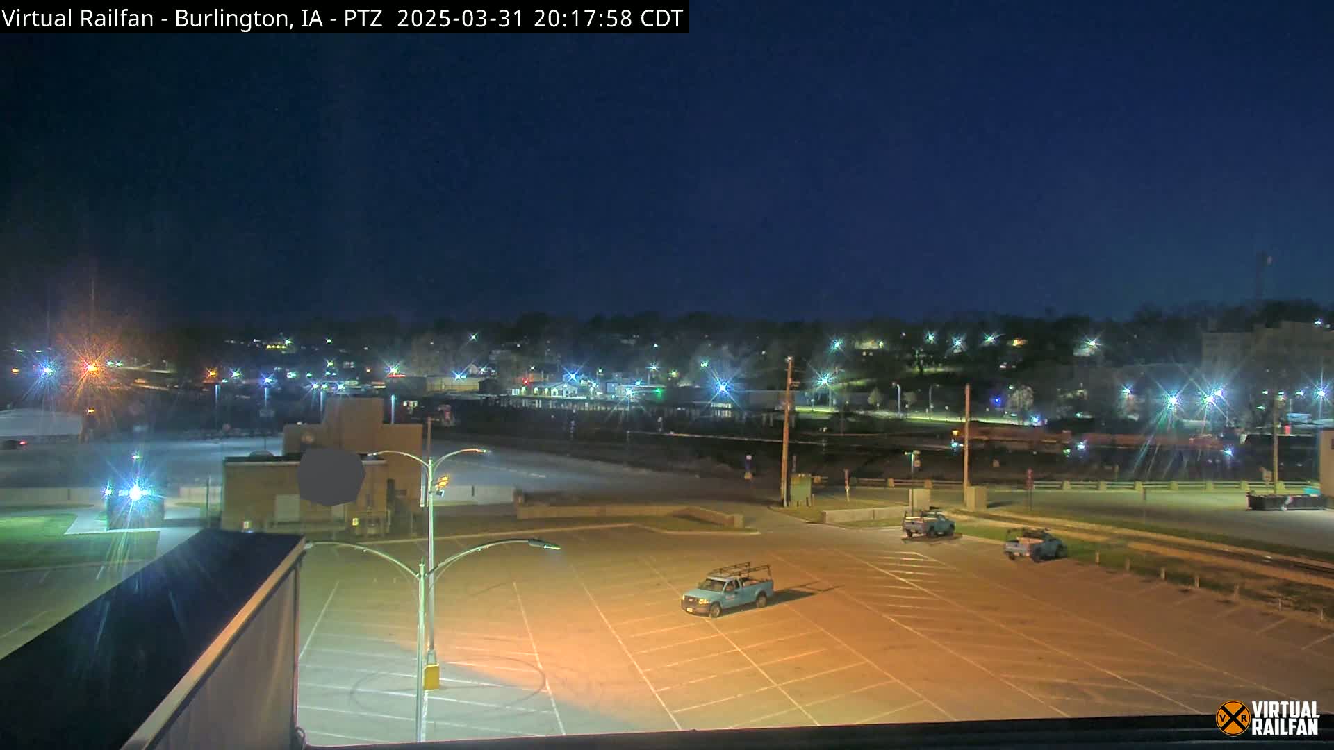 An empty, illuminated parking lot at night is shown, with a few vehicles present and a town in the background under a clear, dark sky.