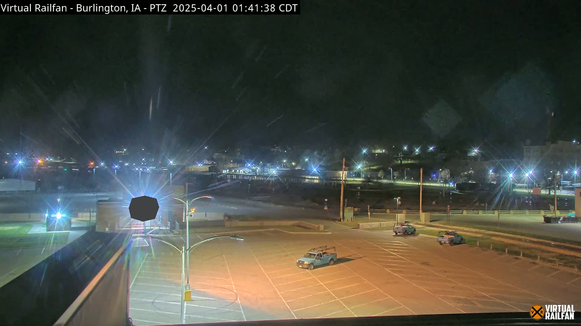A nighttime view of an empty parking lot with a few vehicles, overlooked by a building and a distant town illuminated by streetlights under a clear sky.
